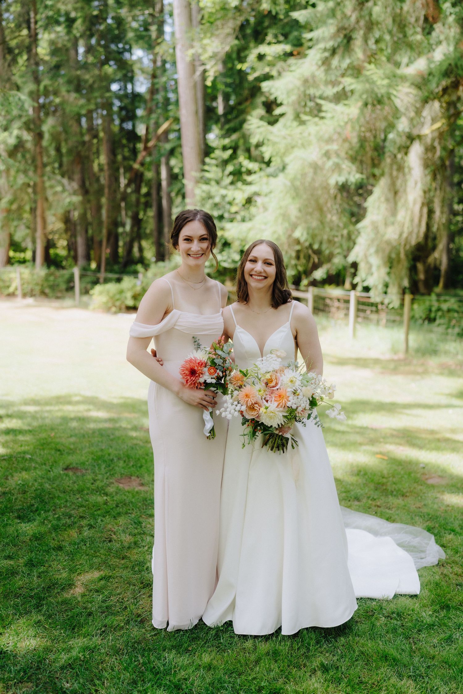 Bridesmaids in white dresses hold colorful bouquets while standing on green grass in a forest setting.