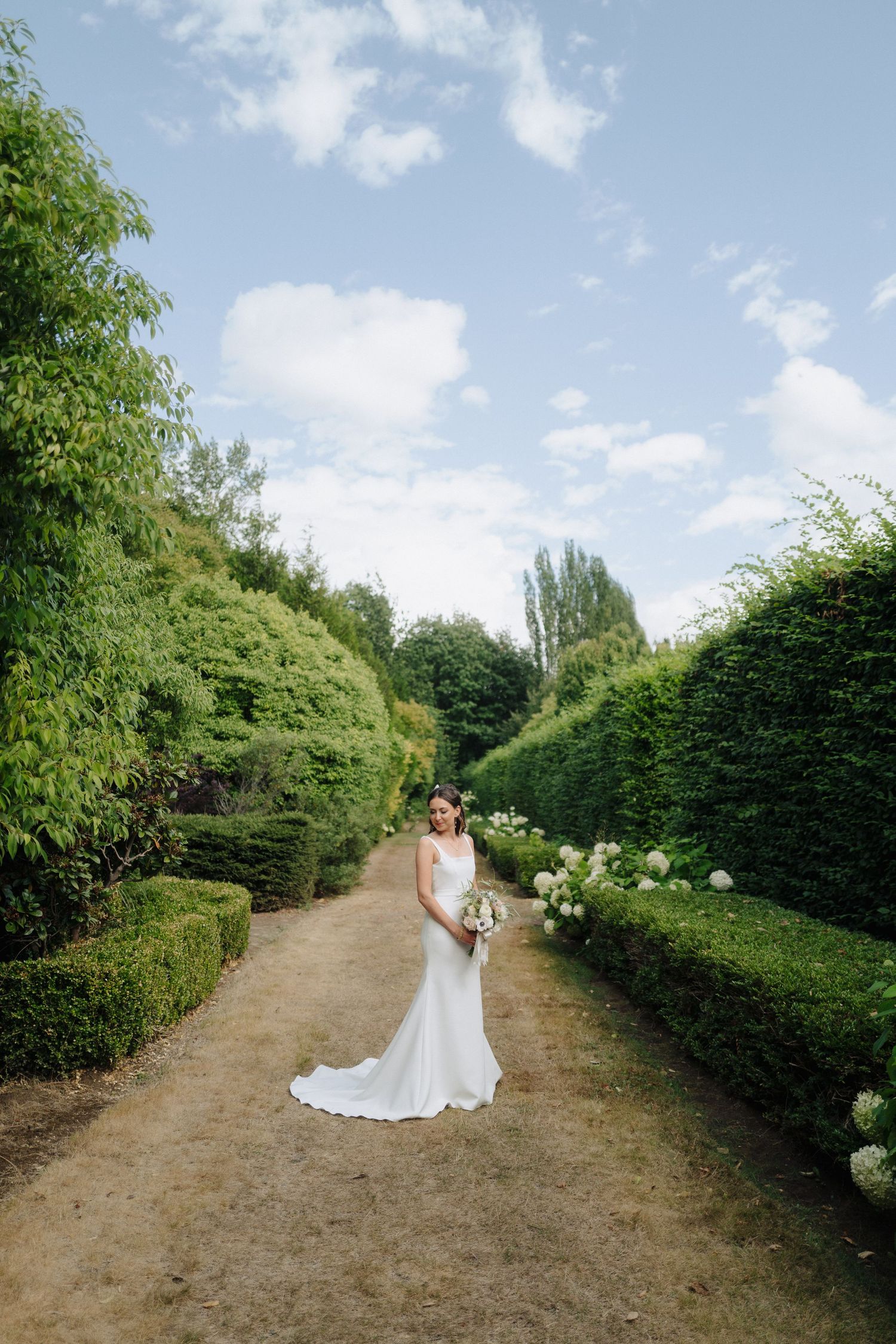 A figure in a white wedding dress stands on a garden path surrounded by manicured hedges.