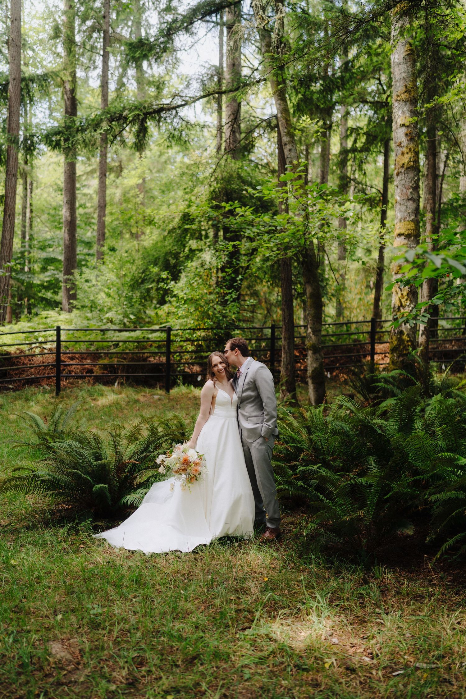 Romantic forest wedding portrait with couple surrounded by lush ferns and evergreen trees.
