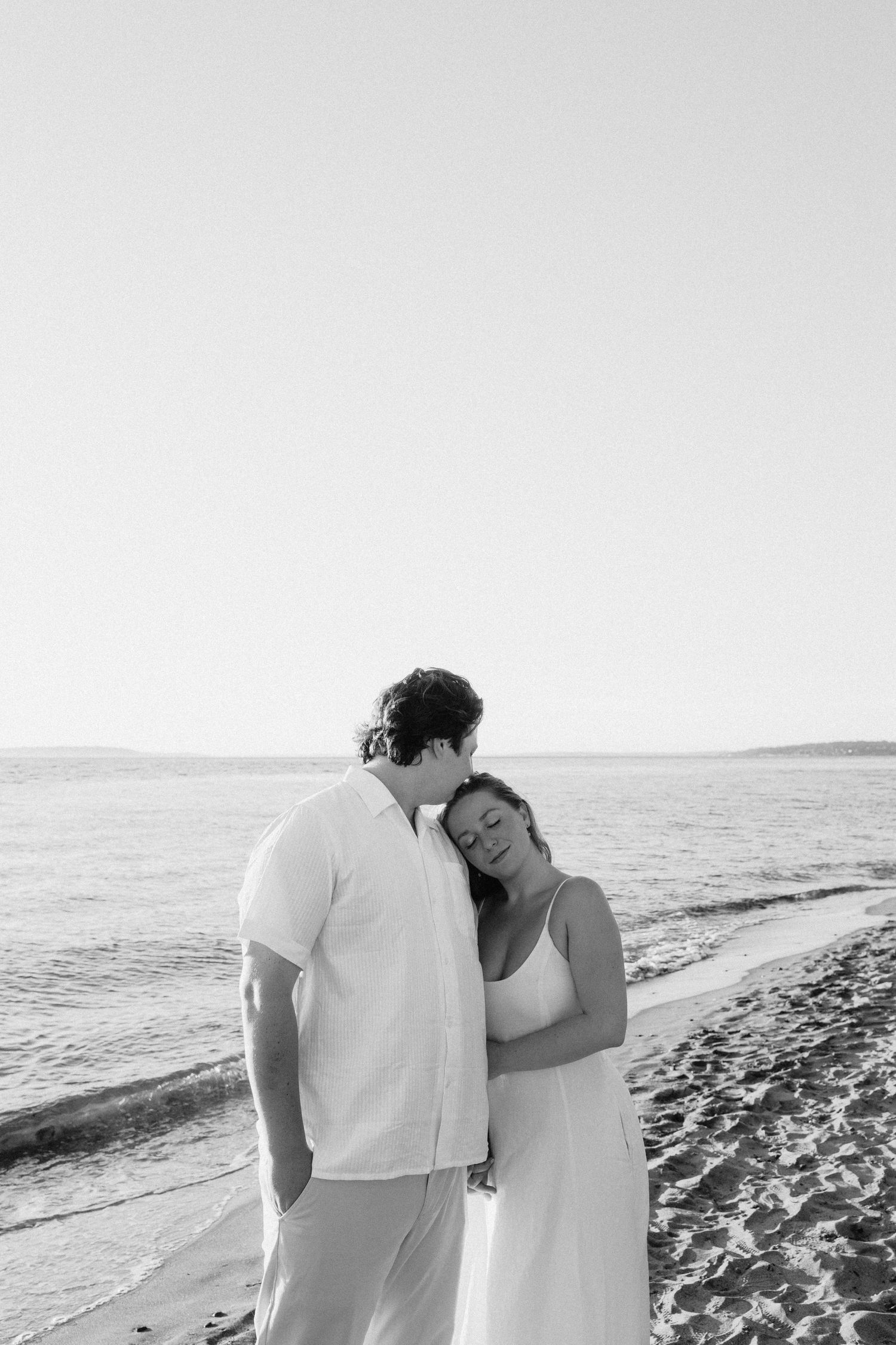 A couple dressed in white embraces tenderly on a beach with the ocean in the background.