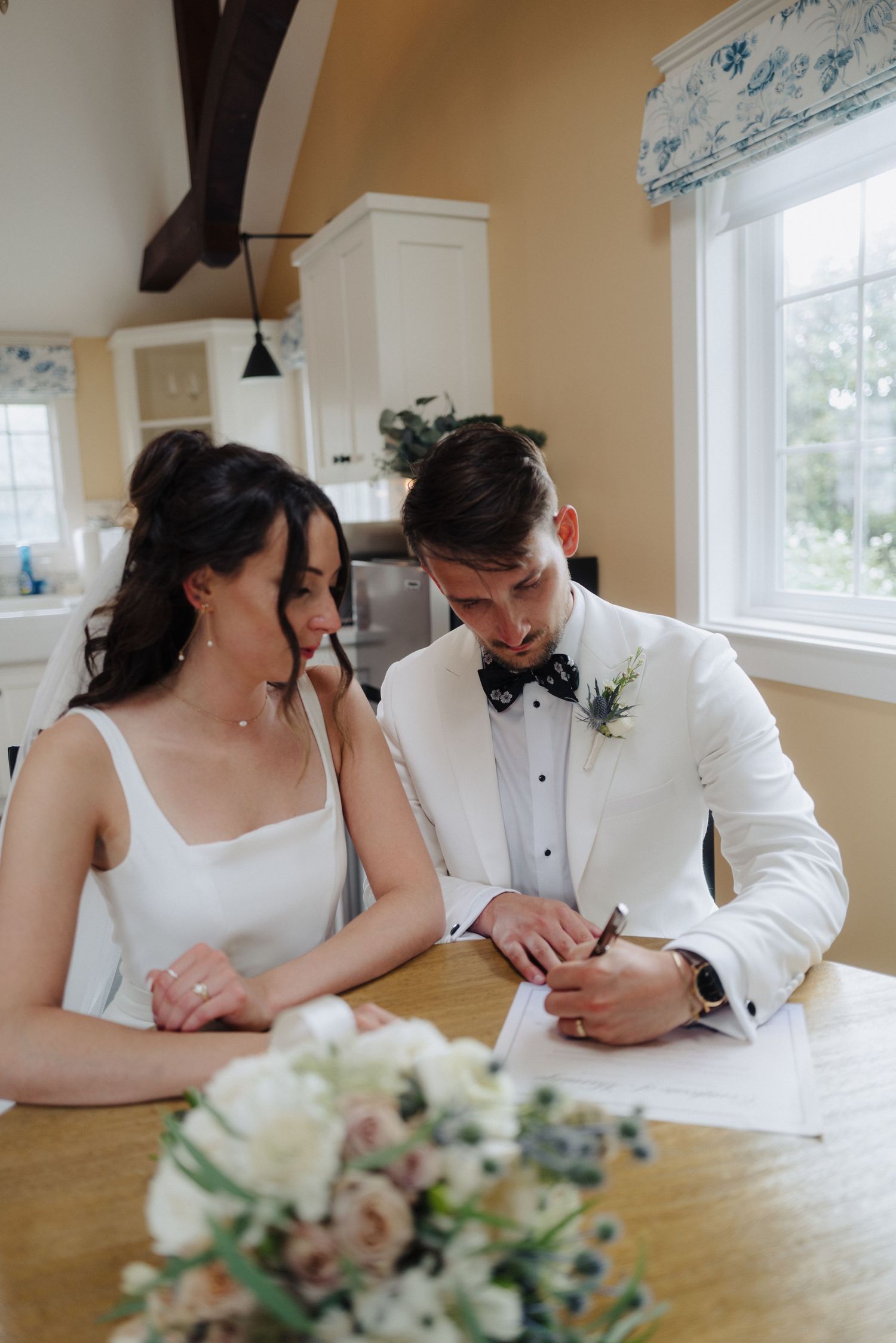 A couple in formal wedding attire sits at a wooden table with floral centerpiece.