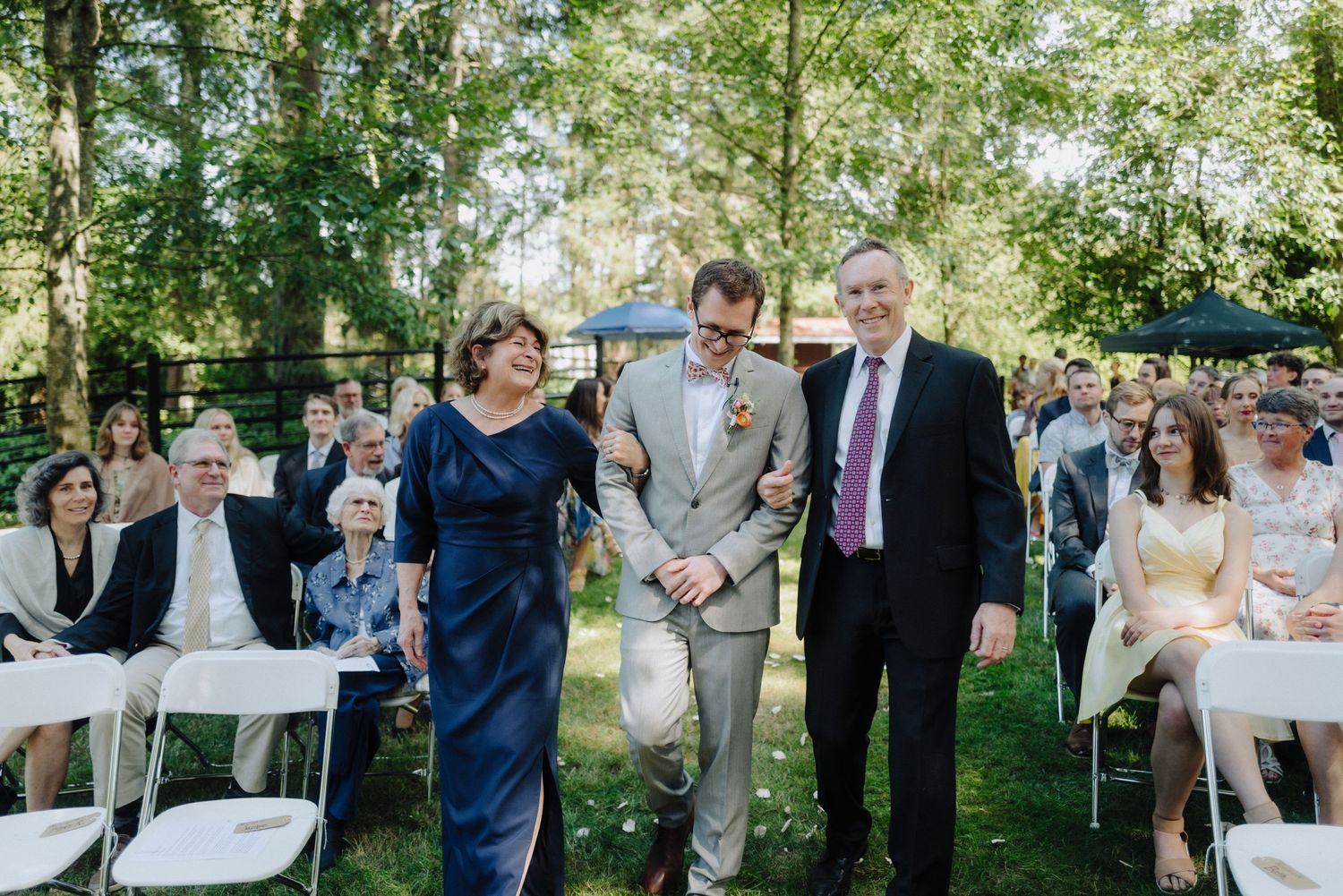Wedding ceremony processional in outdoor garden setting with guests seated on white chairs.