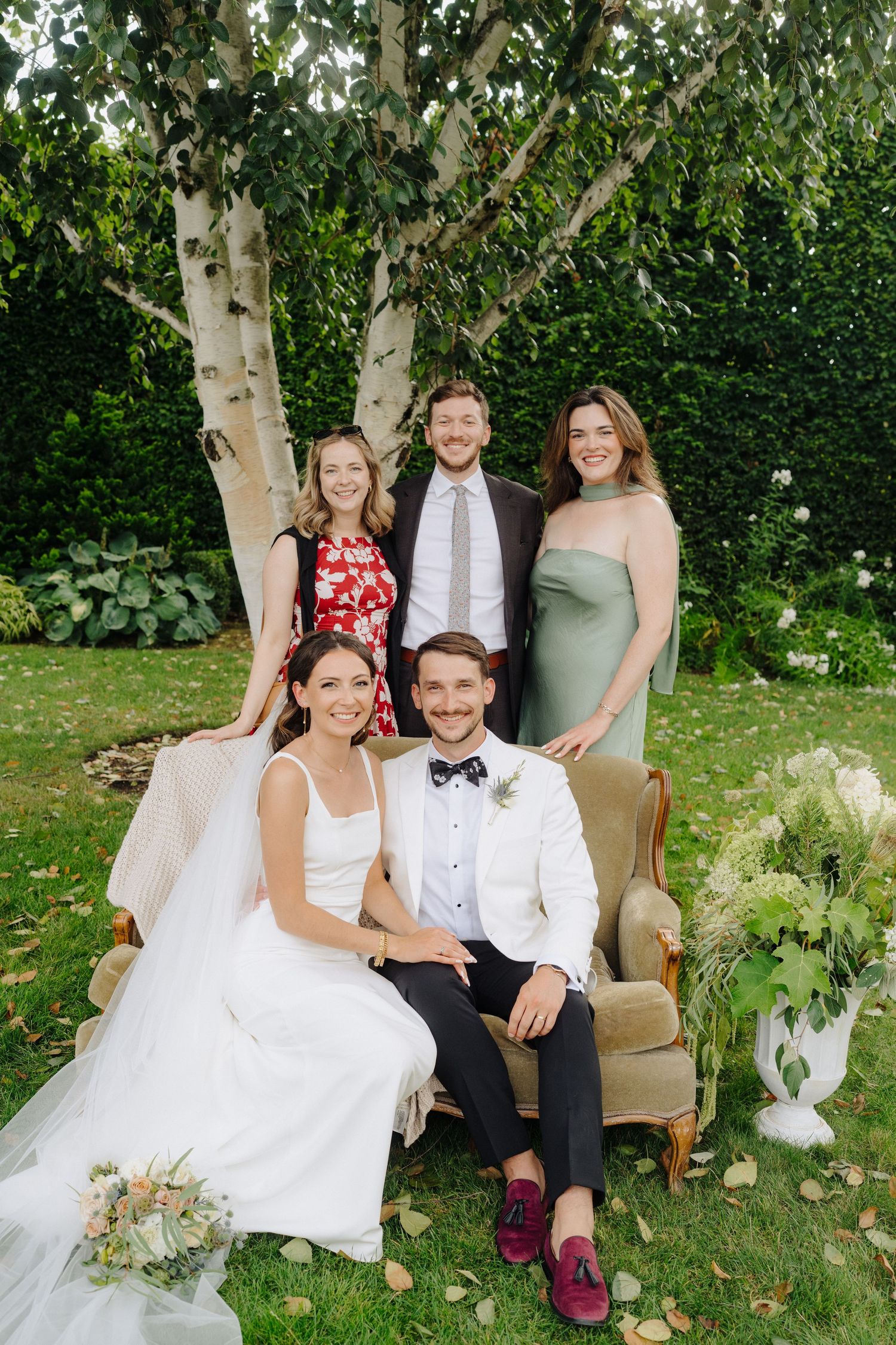 Wedding party sits on vintage chairs in a lush garden setting with elegant decor and floral arrangements.