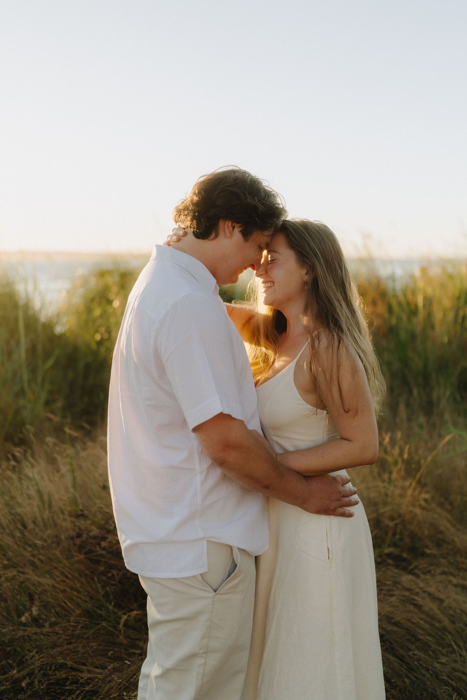 Romantic couple in white outfits embrace during sunset photoshoot in tall grass field.