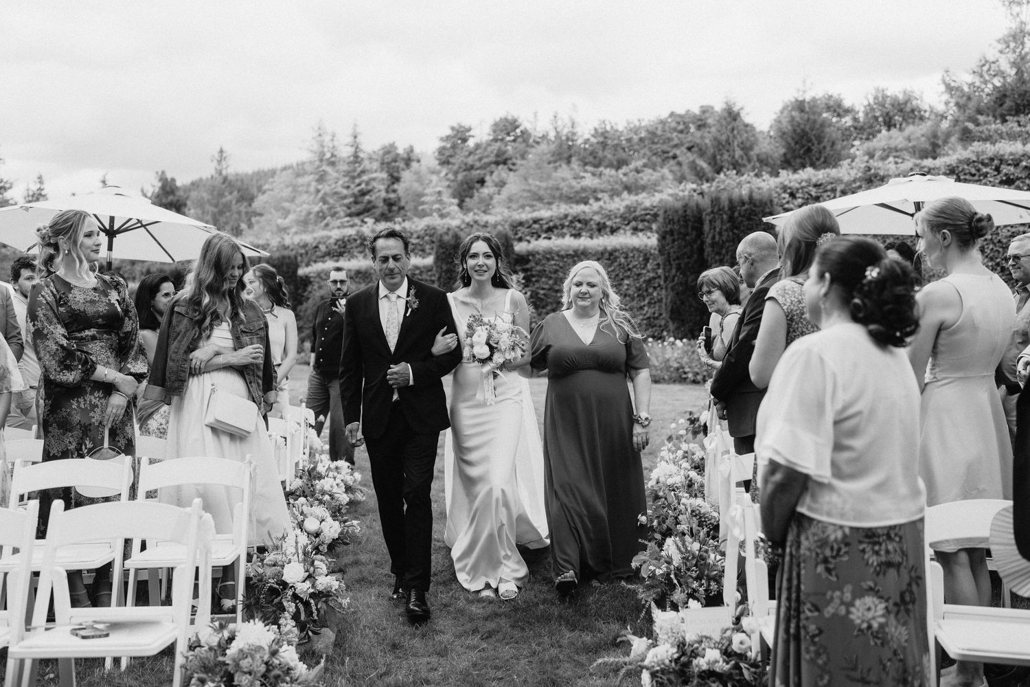 Black and white wedding ceremony processional with guests seated in outdoor garden setting.