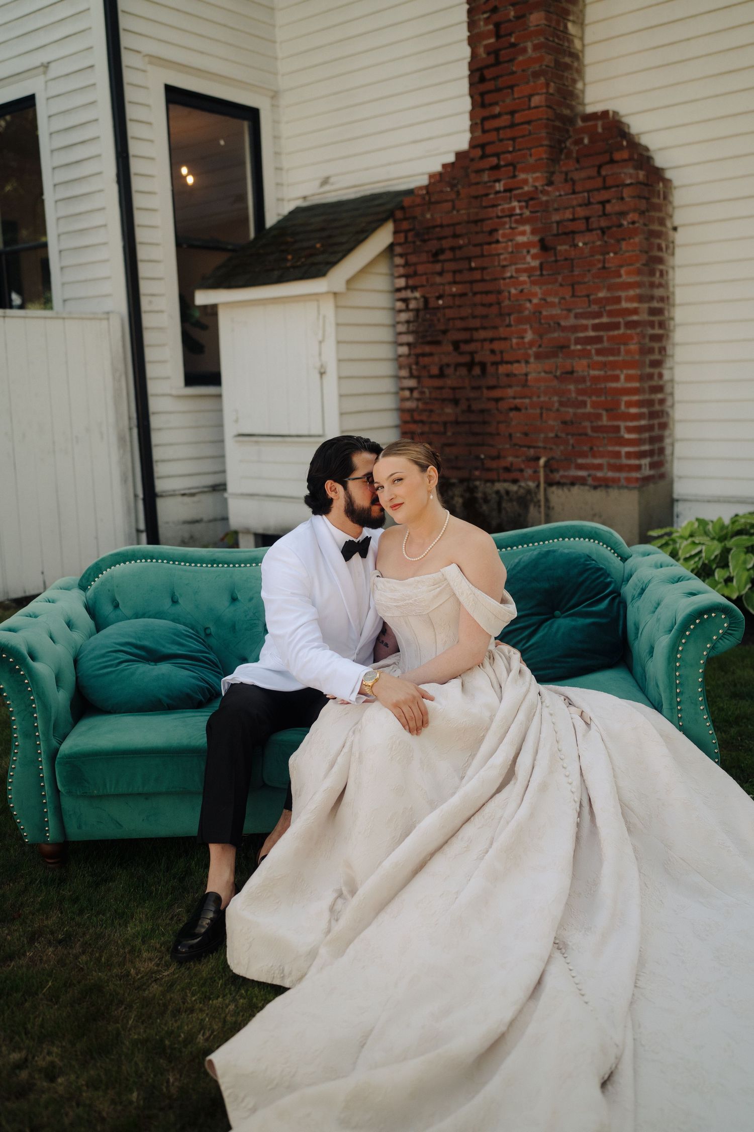 Couple in wedding attire lounges on emerald green velvet couch against white house with brick chimney.