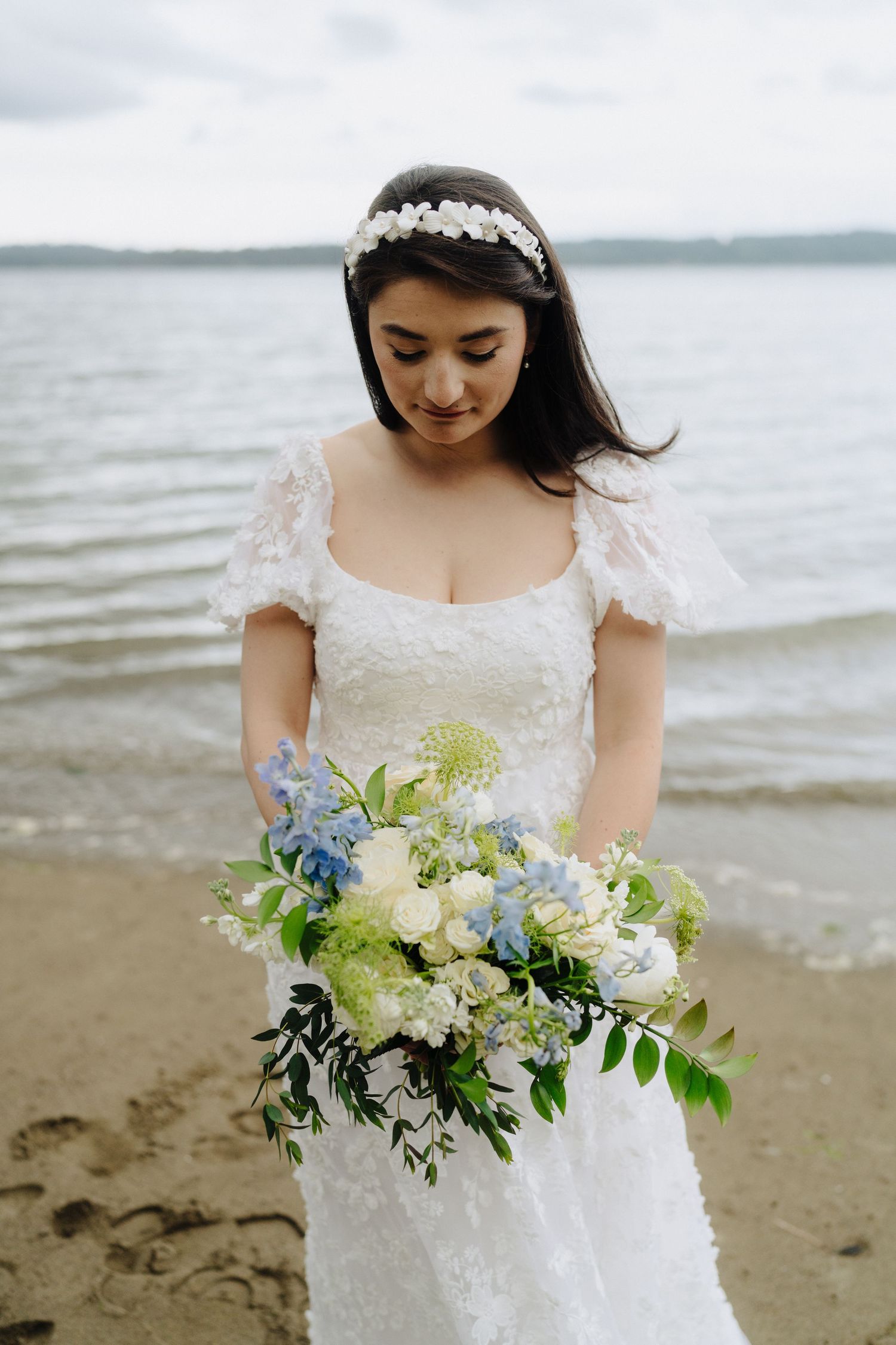 A bride in a lace wedding dress holds a delicate bouquet of white and blue flowers by the waterfront.