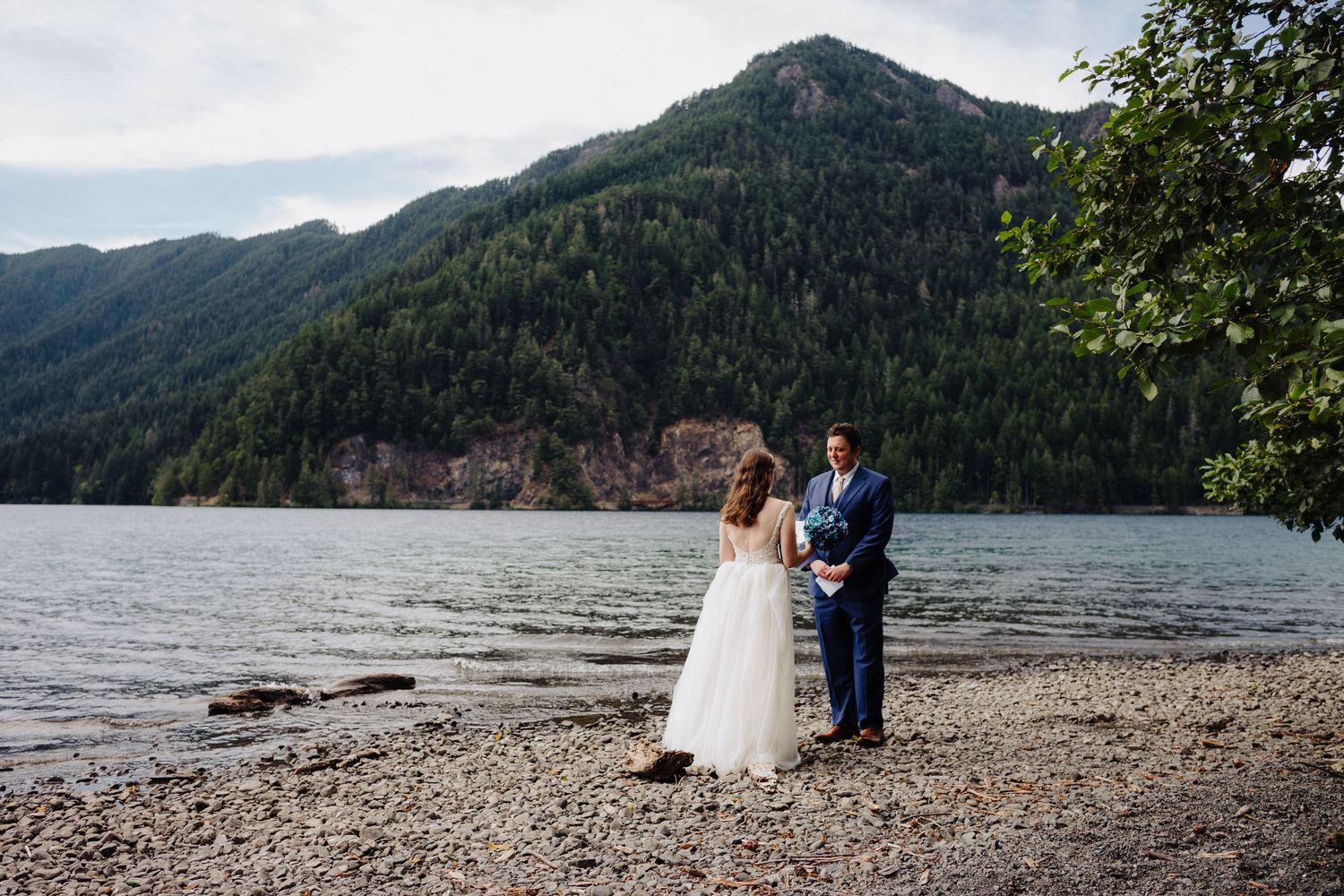 A couple poses for wedding photos on a rocky beach with forested mountains in the background.