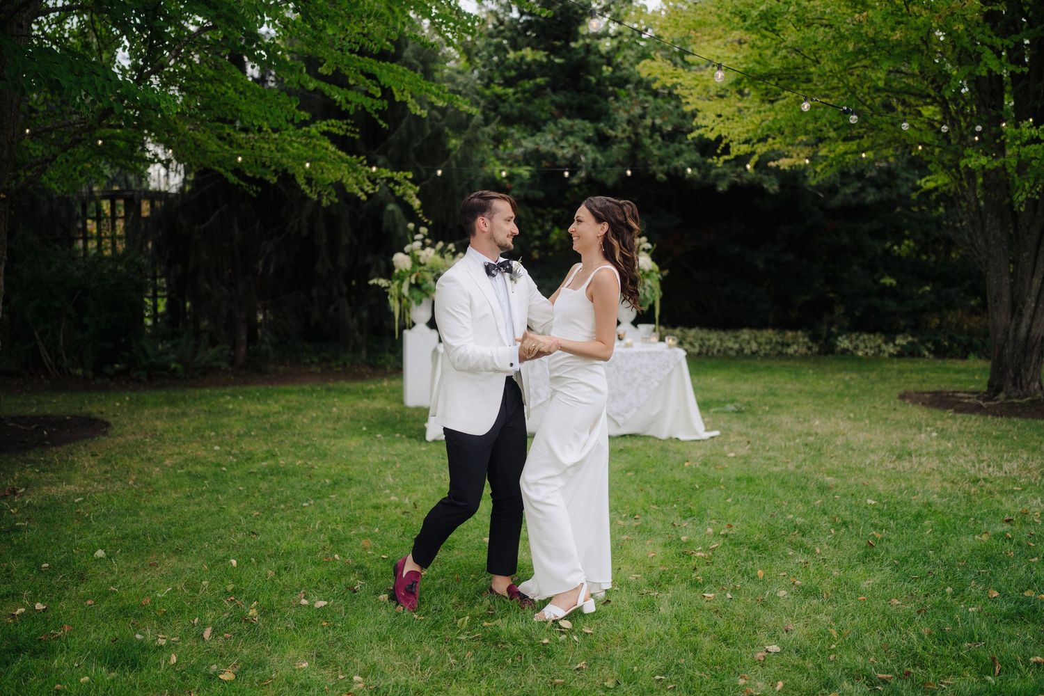 Wedding couples dance on a sunlit lawn surrounded by green trees during an outdoor reception.