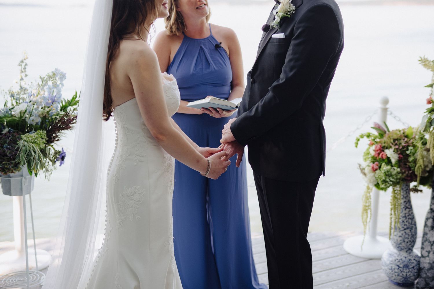 Close-up of couple holding hands during waterfront wedding ceremony with officiant in blue.