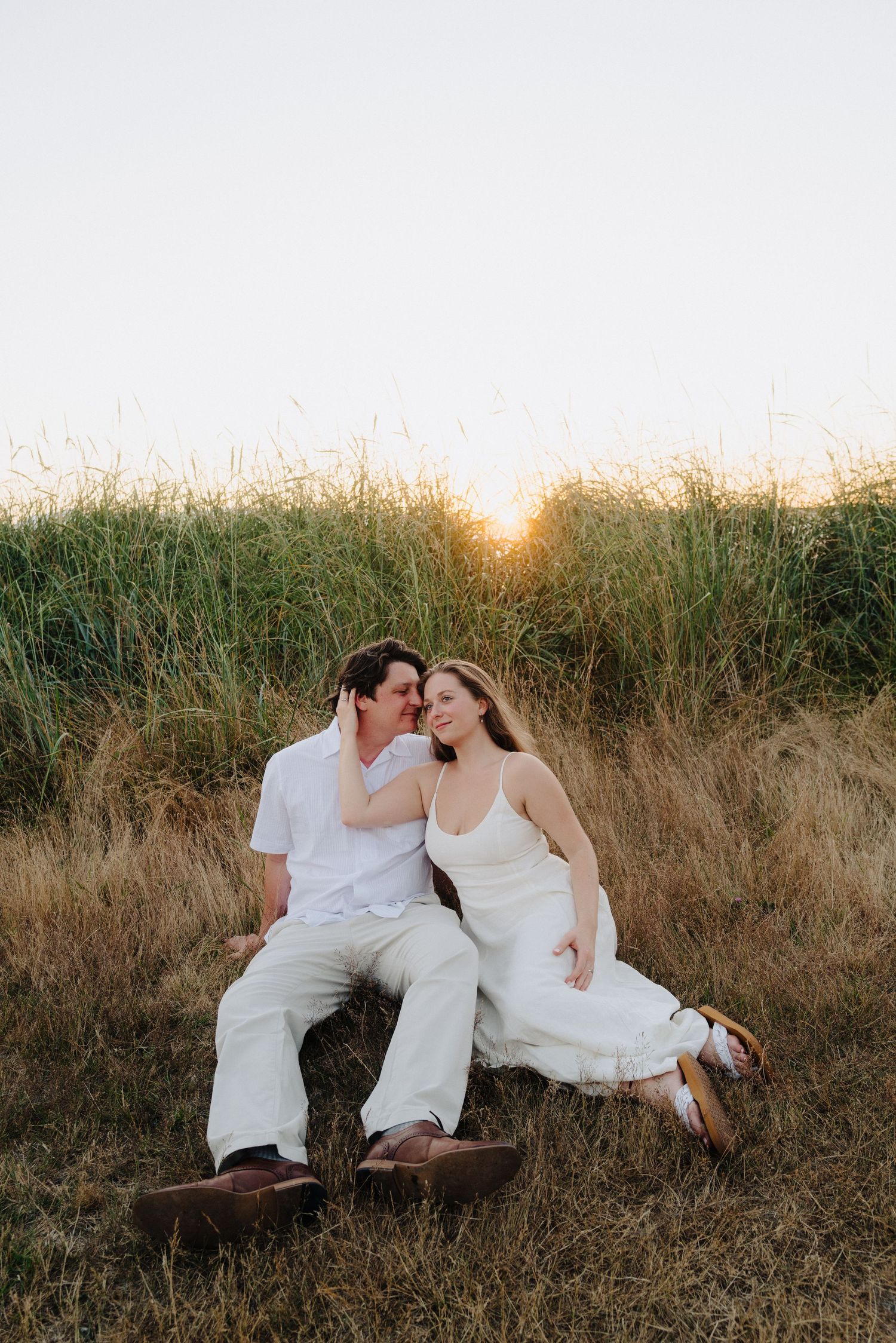 A couple in white outfits embraces while sitting in a golden field during sunset.