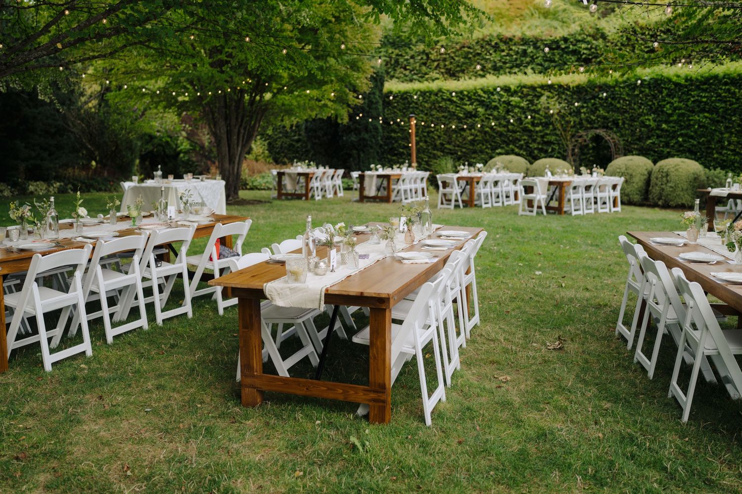 Outdoor wedding reception setup with wooden farm tables and white chairs on manicured lawn.