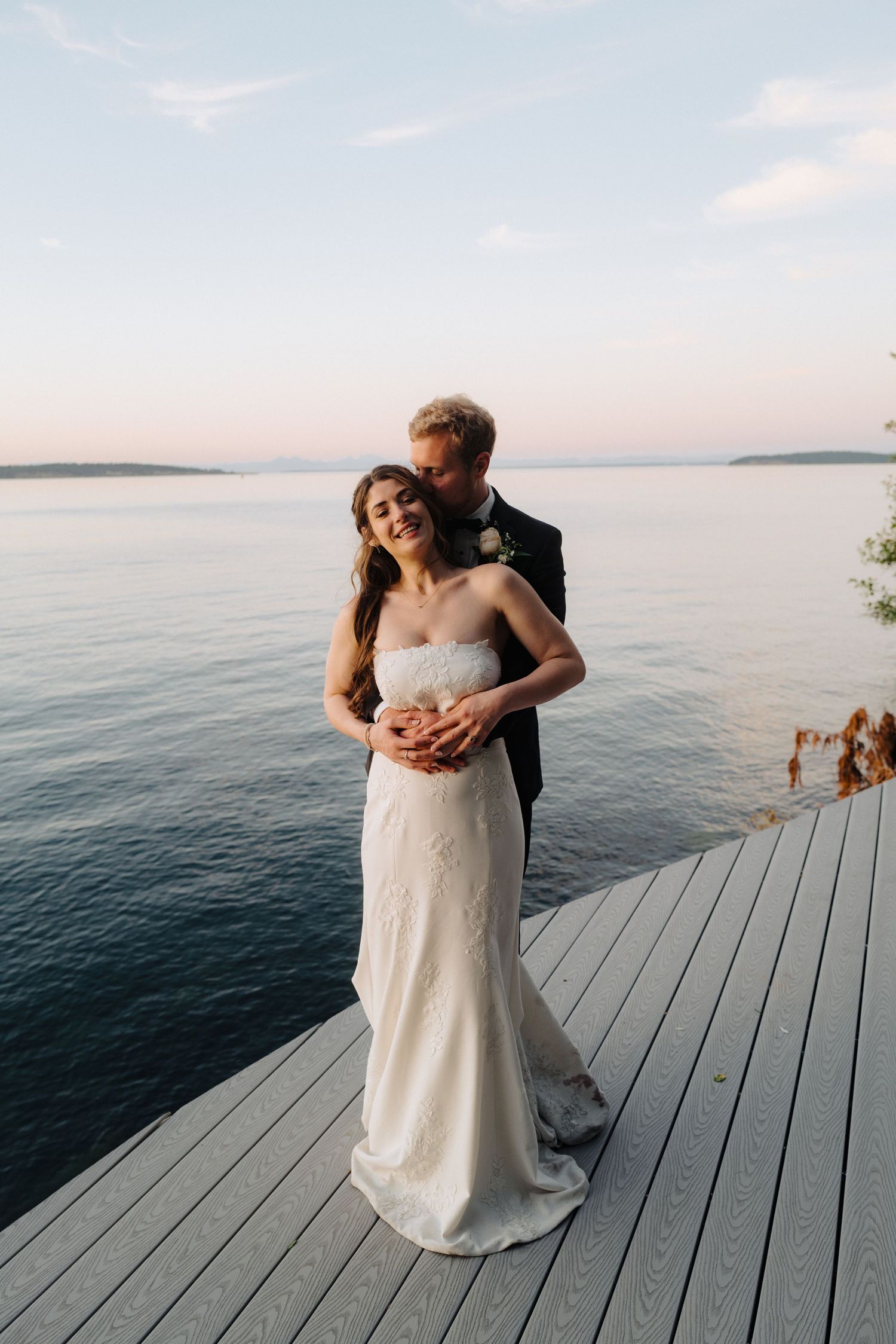 Couple embraces on a wooden deck overlooking a serene lake at sunset.