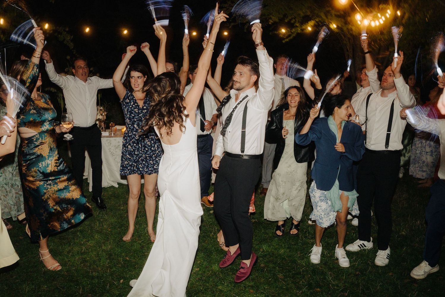 Wedding reception guests dancing under string lights at an outdoor nighttime celebration.