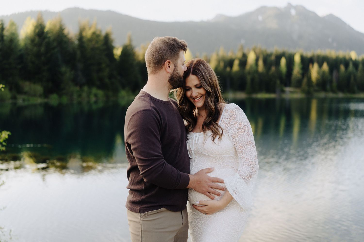 A couple embraces by a tranquil mountain lake surrounded by evergreen forest and snow-capped peaks.
