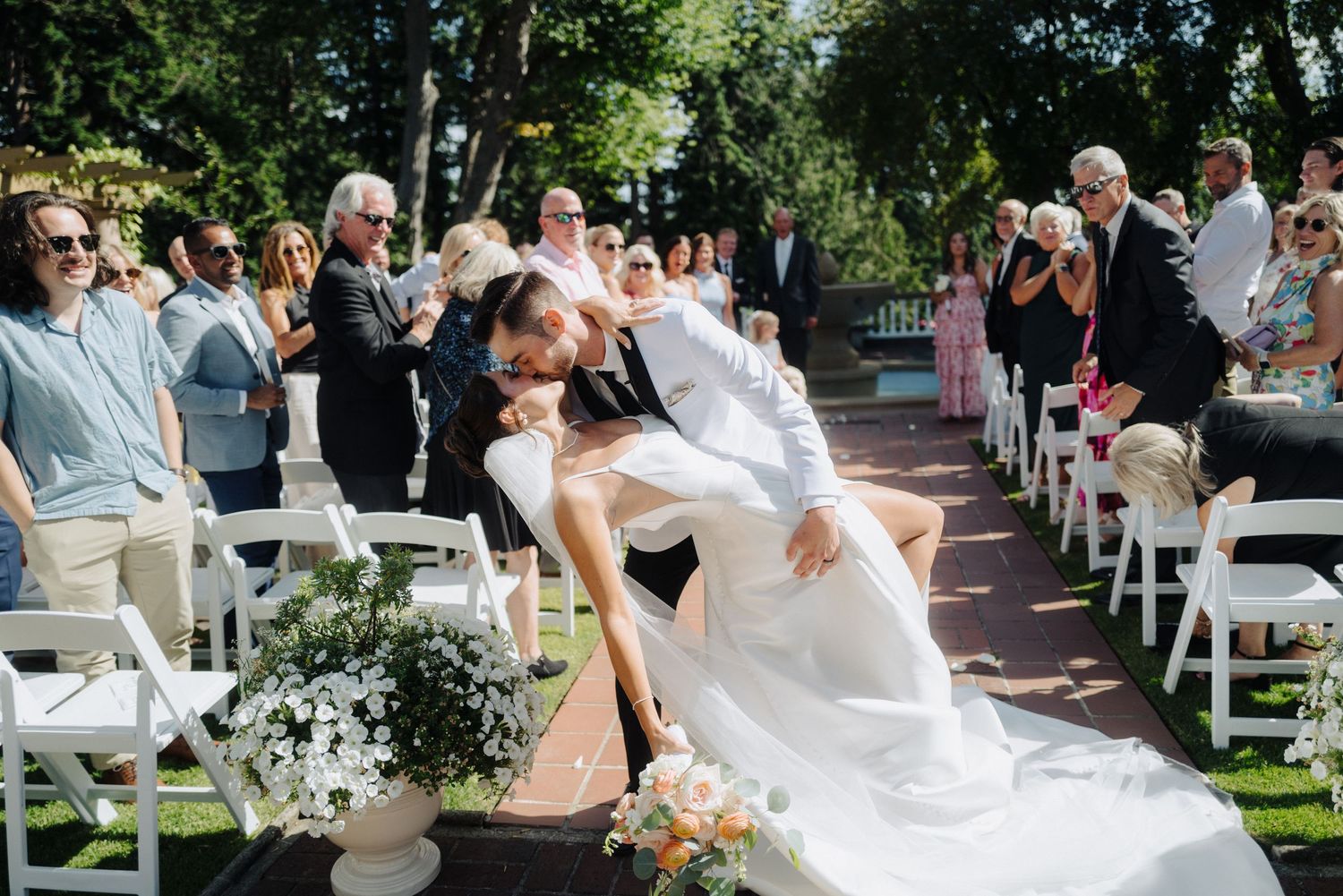 Wide shot of a wedding ceremony in progress with guests seated in white chairs under trees.
