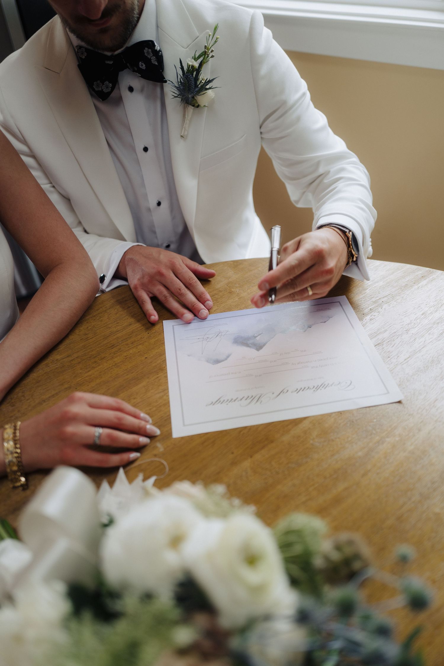 A person in a white tuxedo jacket signs a wedding document with white floral arrangements on the table.