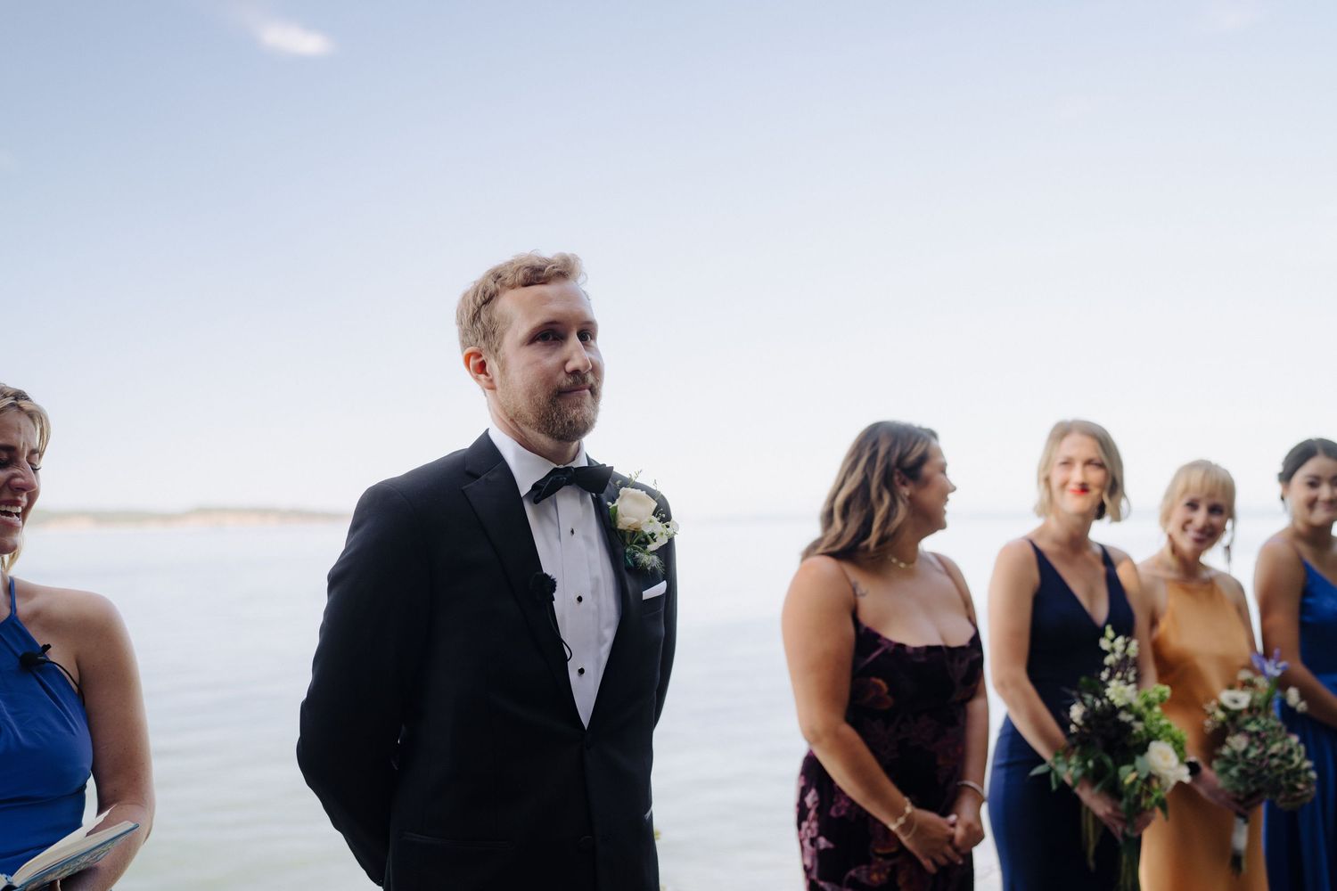 Wedding party stands in line on a deck overlooking a scenic water view on a sunny day.