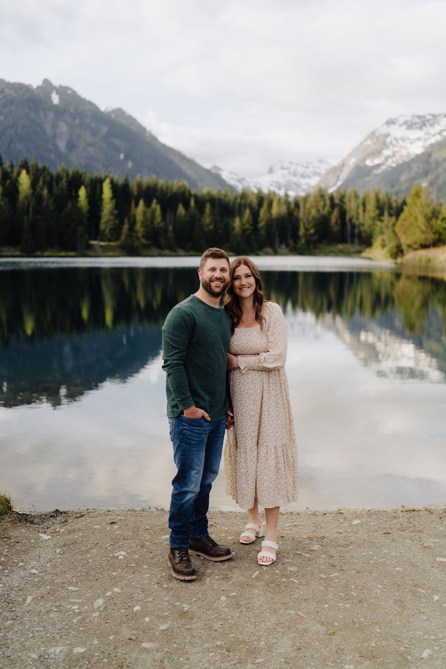 A couple poses by a serene mountain lake surrounded by evergreen forest and snow-capped peaks.