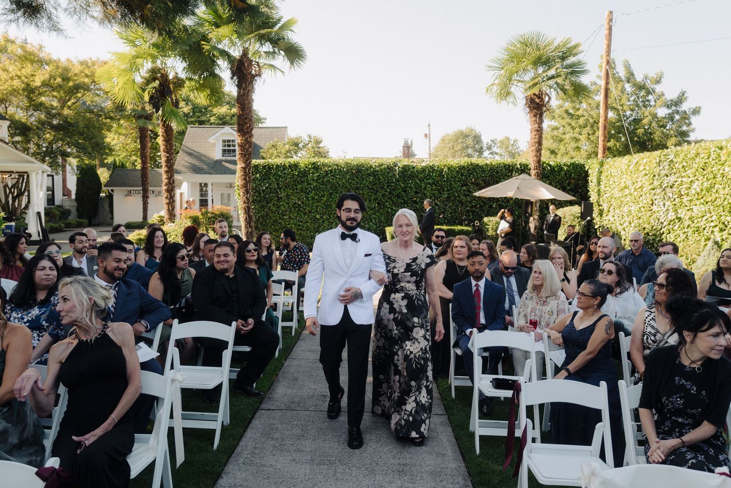 Outdoor wedding ceremony with guests seated in white chairs under trees.