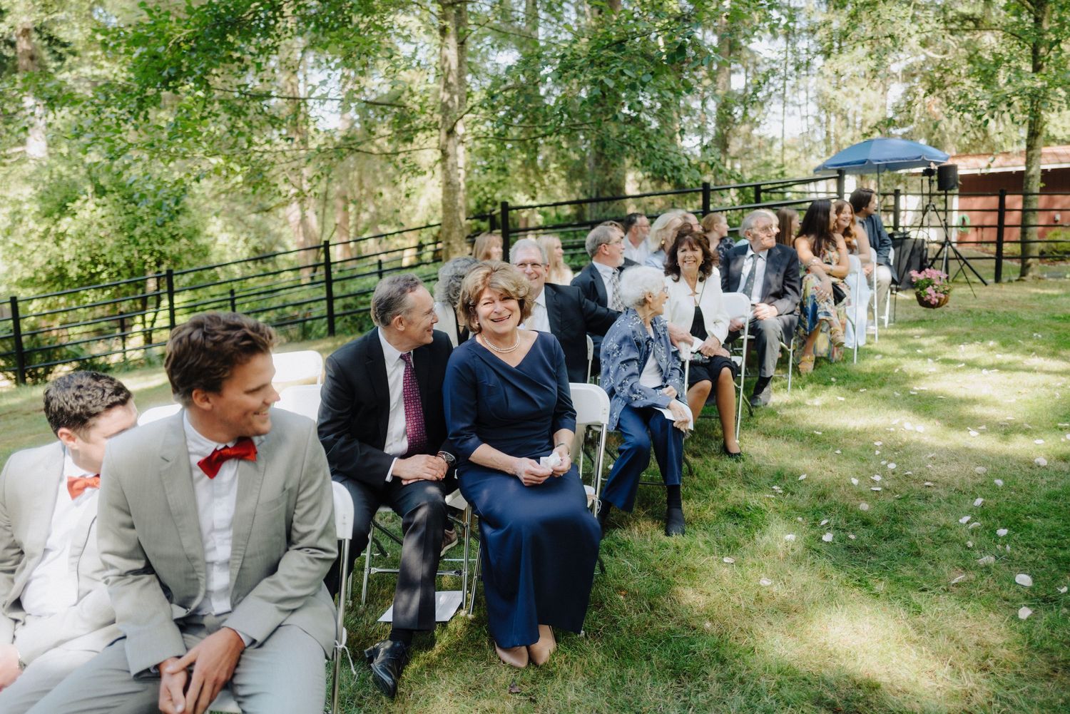 Wedding guests seated in rows of white chairs during outdoor forest ceremony.