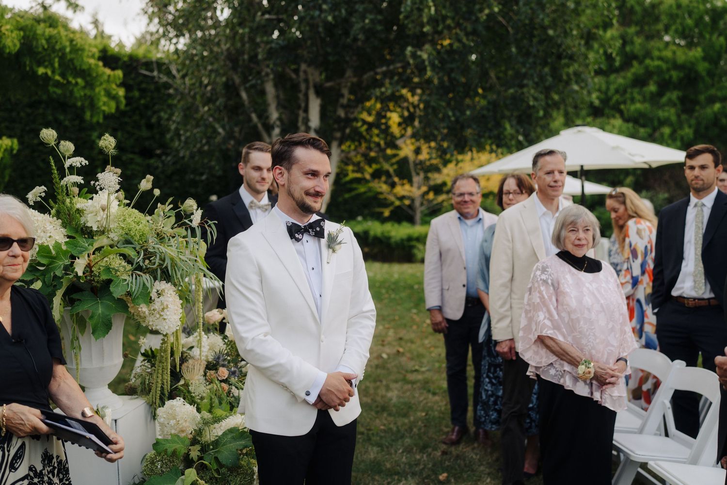 Wedding guests gather outdoors under trees for a garden ceremony celebration.