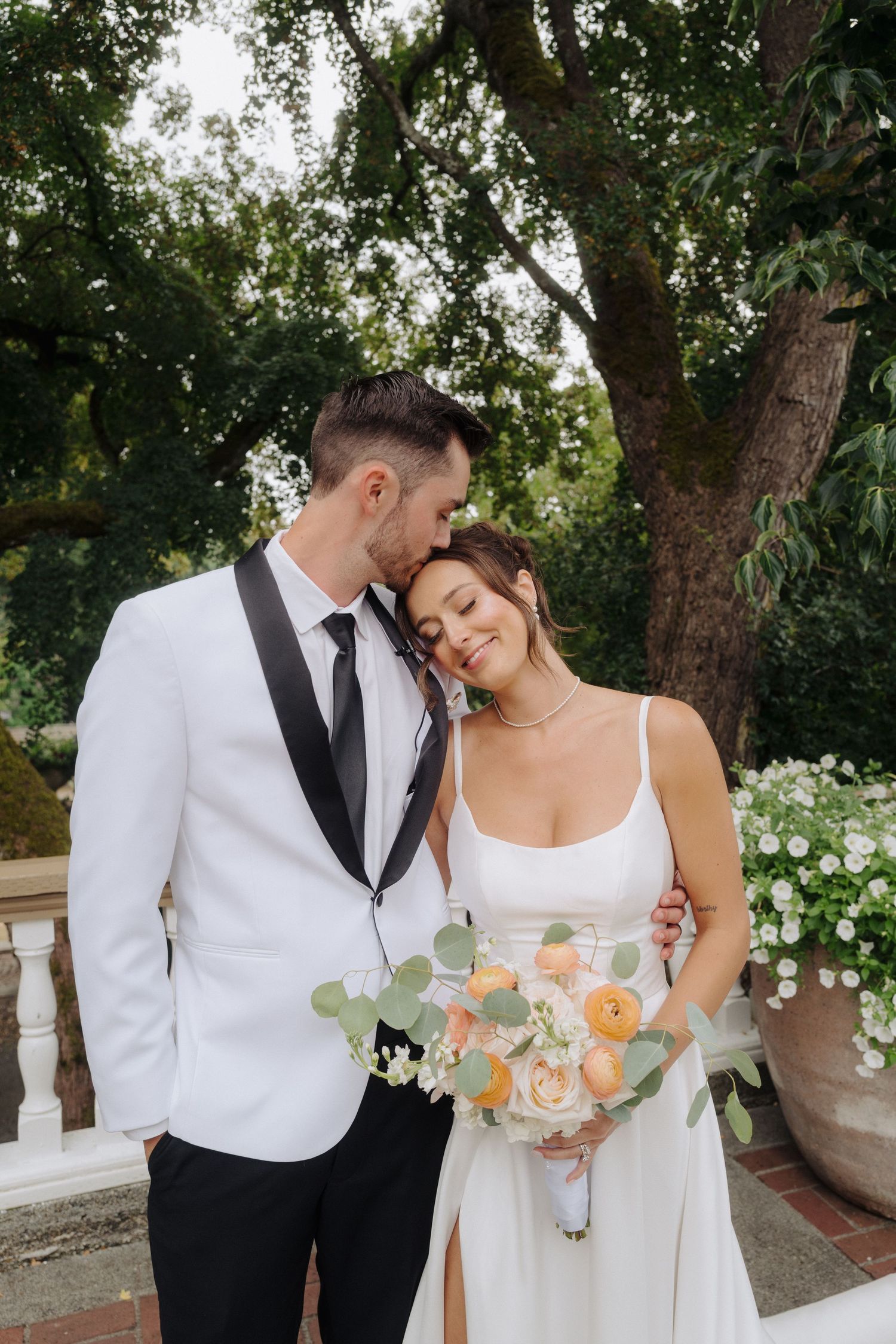 Newlyweds share tender moment in garden setting with peach rose bouquet.
