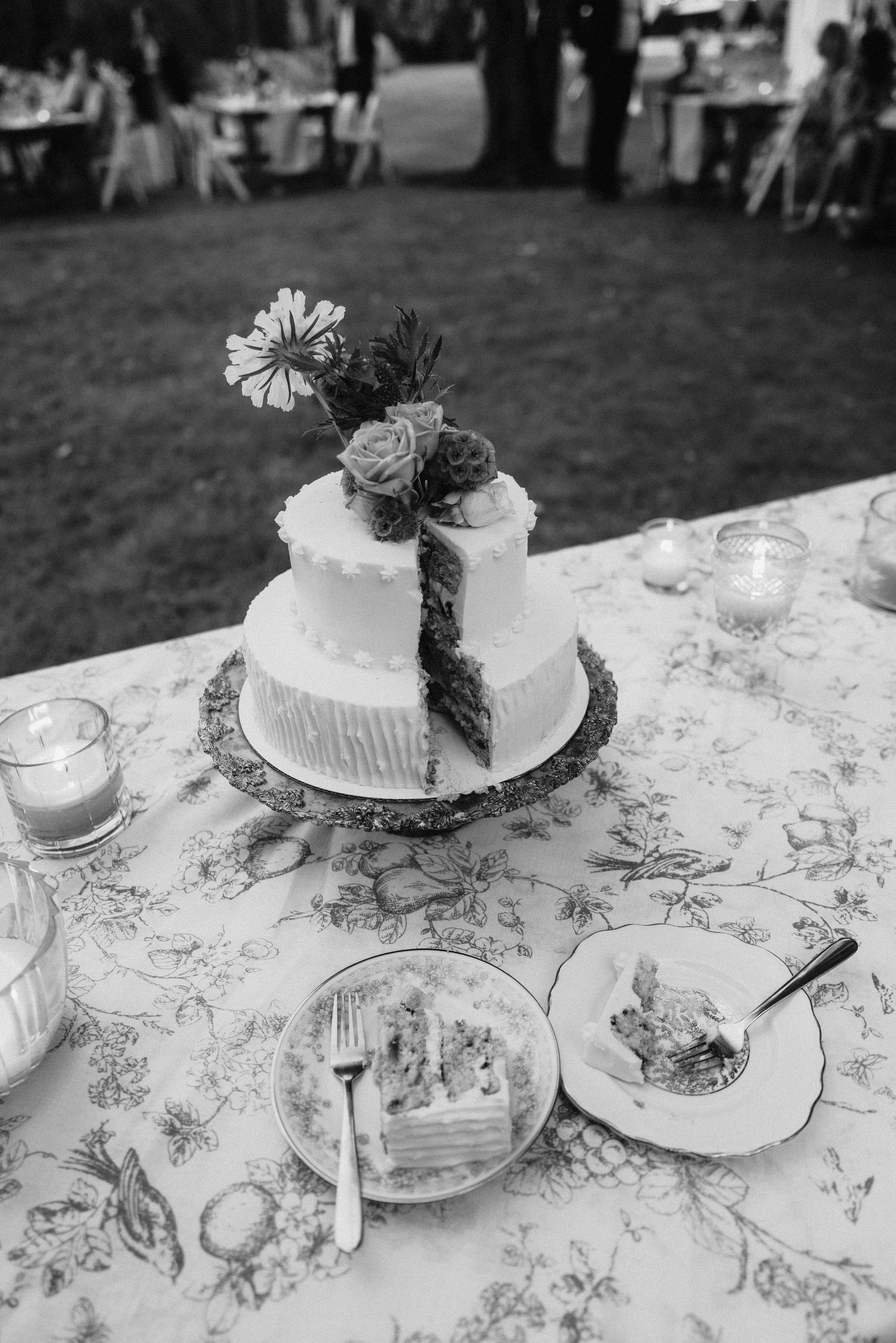 White wedding cake with floral decorations displayed on patterned tablecloth.