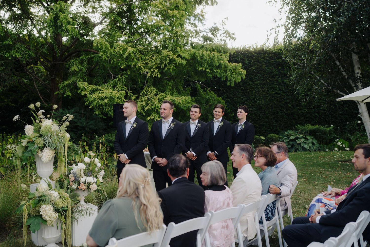 Wedding ceremony setup in garden with guests seated and tall floral arrangements framing the aisle.
