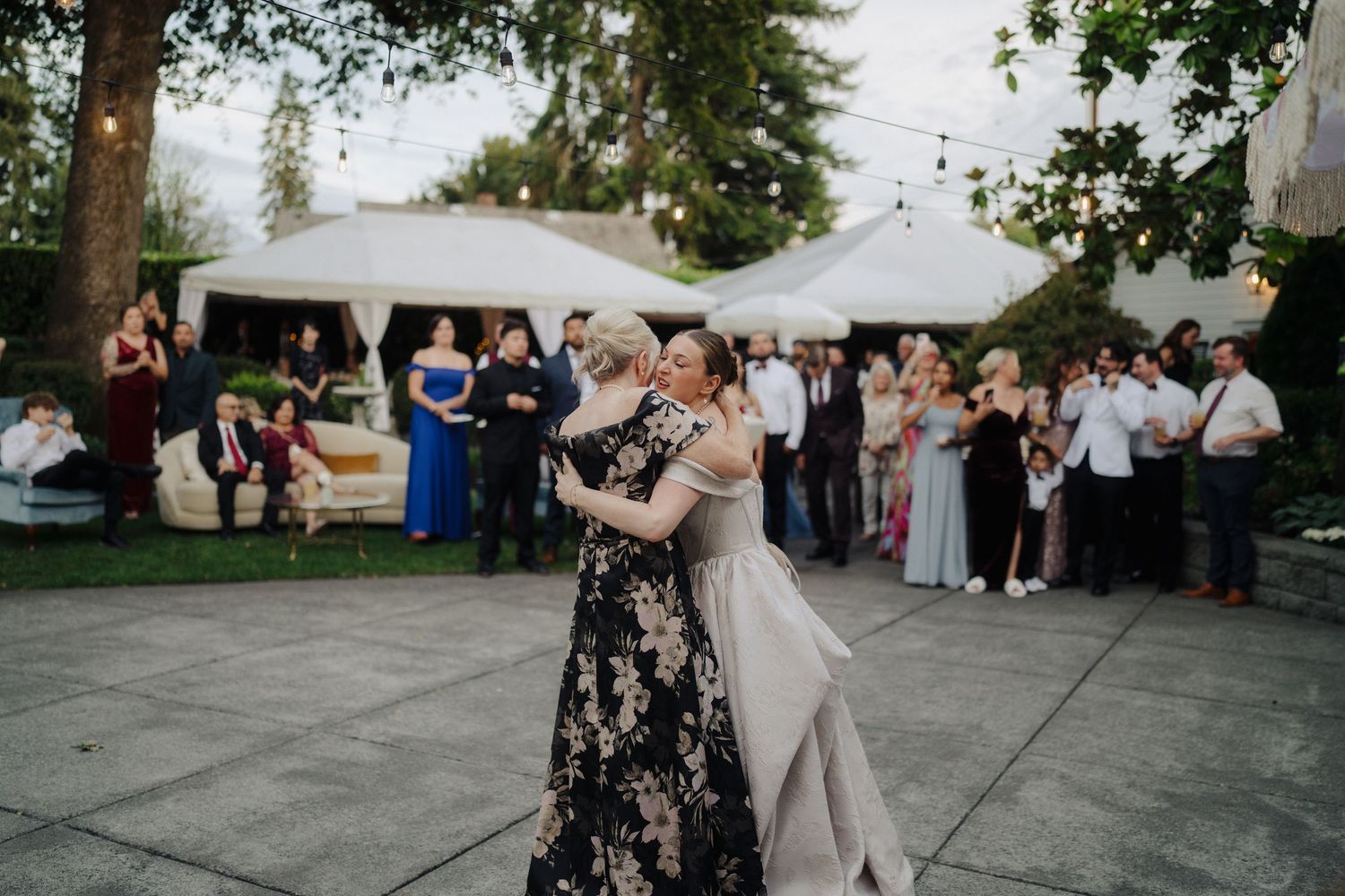 Outdoor wedding reception with guests mingling under white tent and tree canopy.