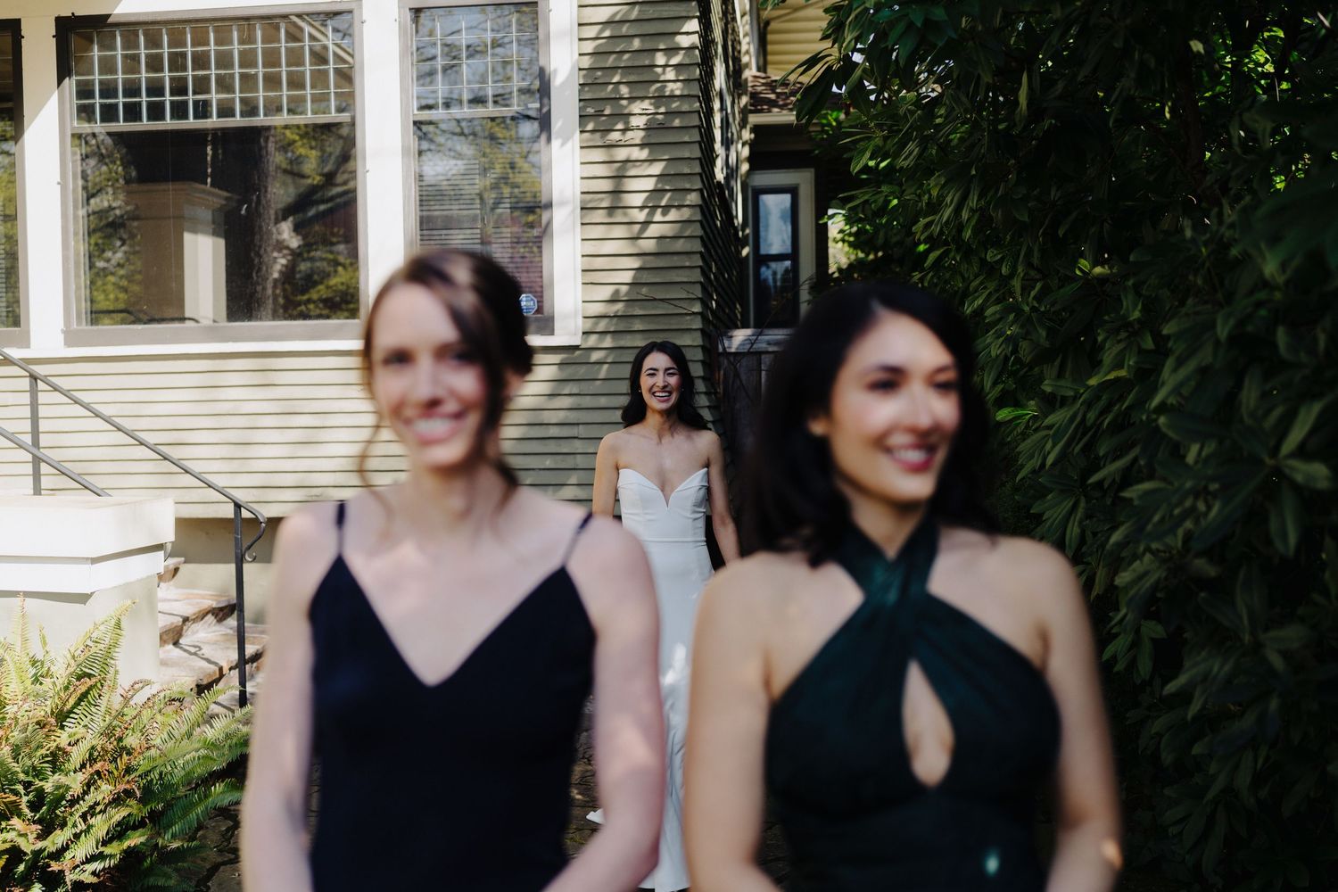 Bridesmaids in elegant black dresses walk outside a white house surrounded by greenery.