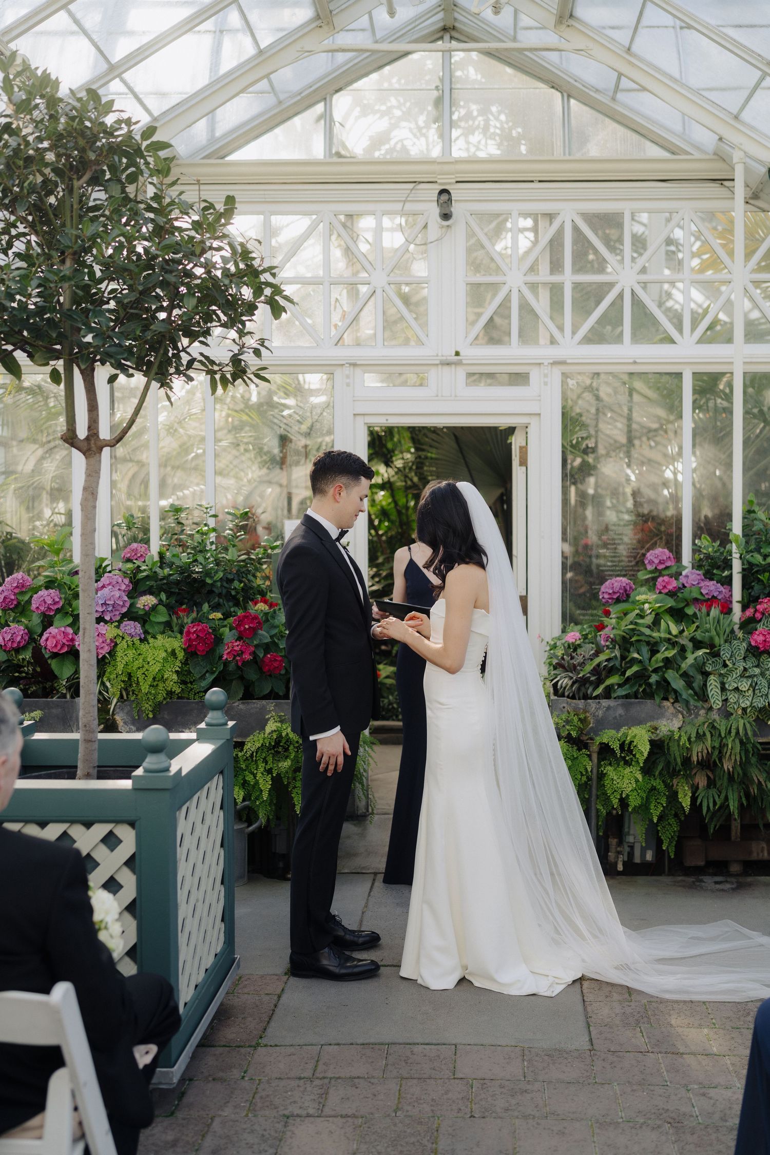 A wedding ceremony in a greenhouse filled with pink flowers and lush greenery.