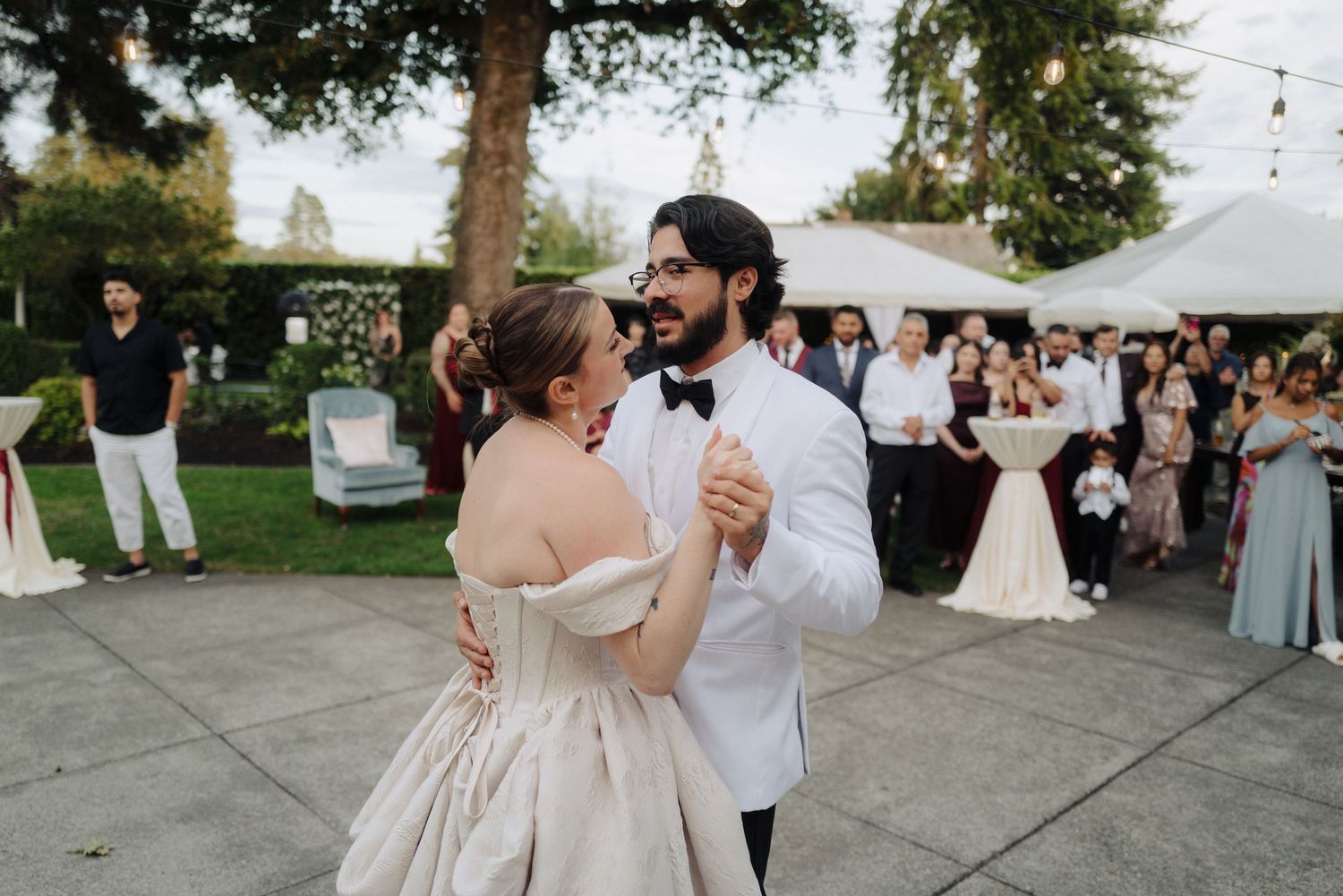 A couple shares their first dance at an outdoor wedding reception surrounded by guests and trees.