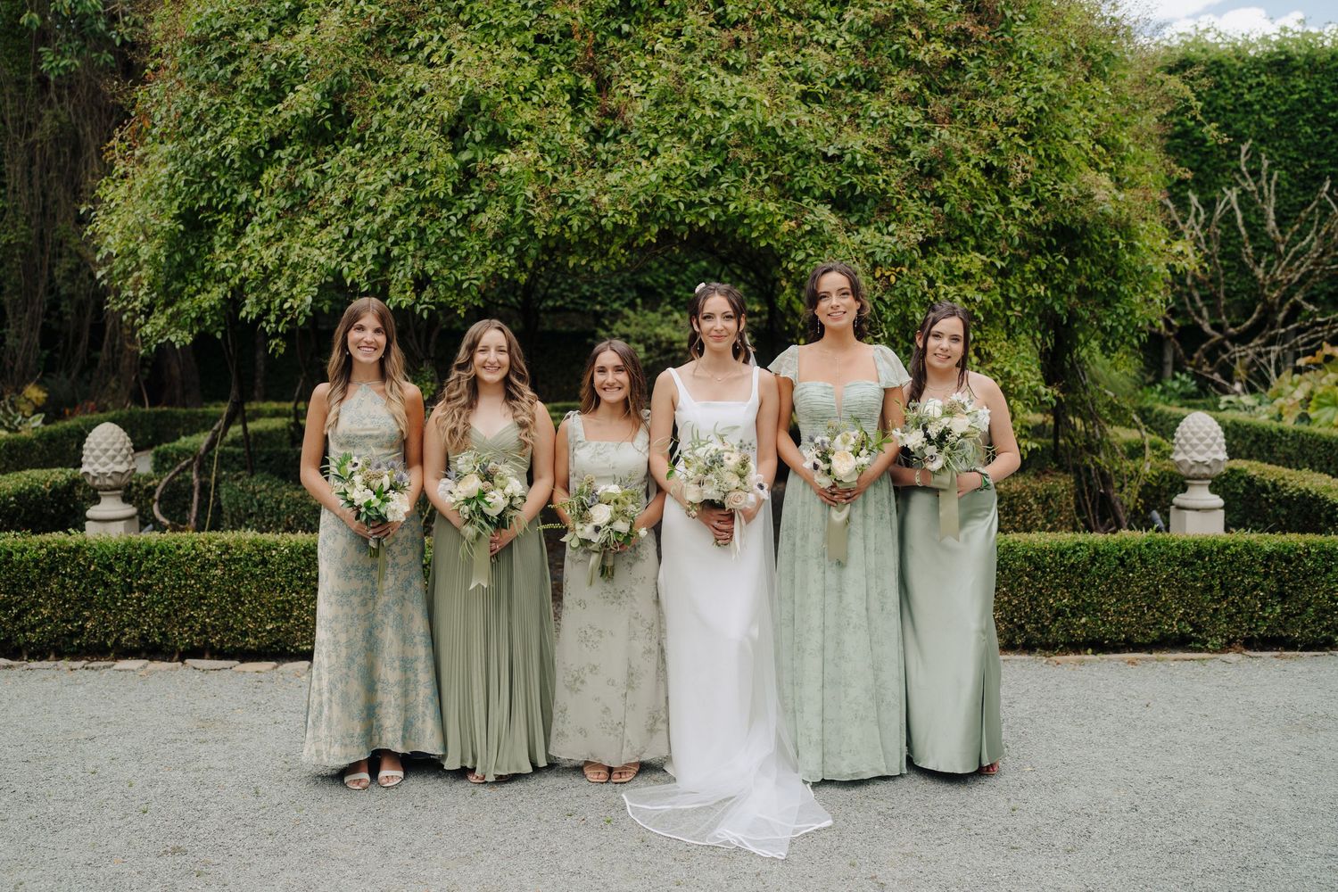 Bridesmaids in sage green dresses pose with bride on garden grounds.