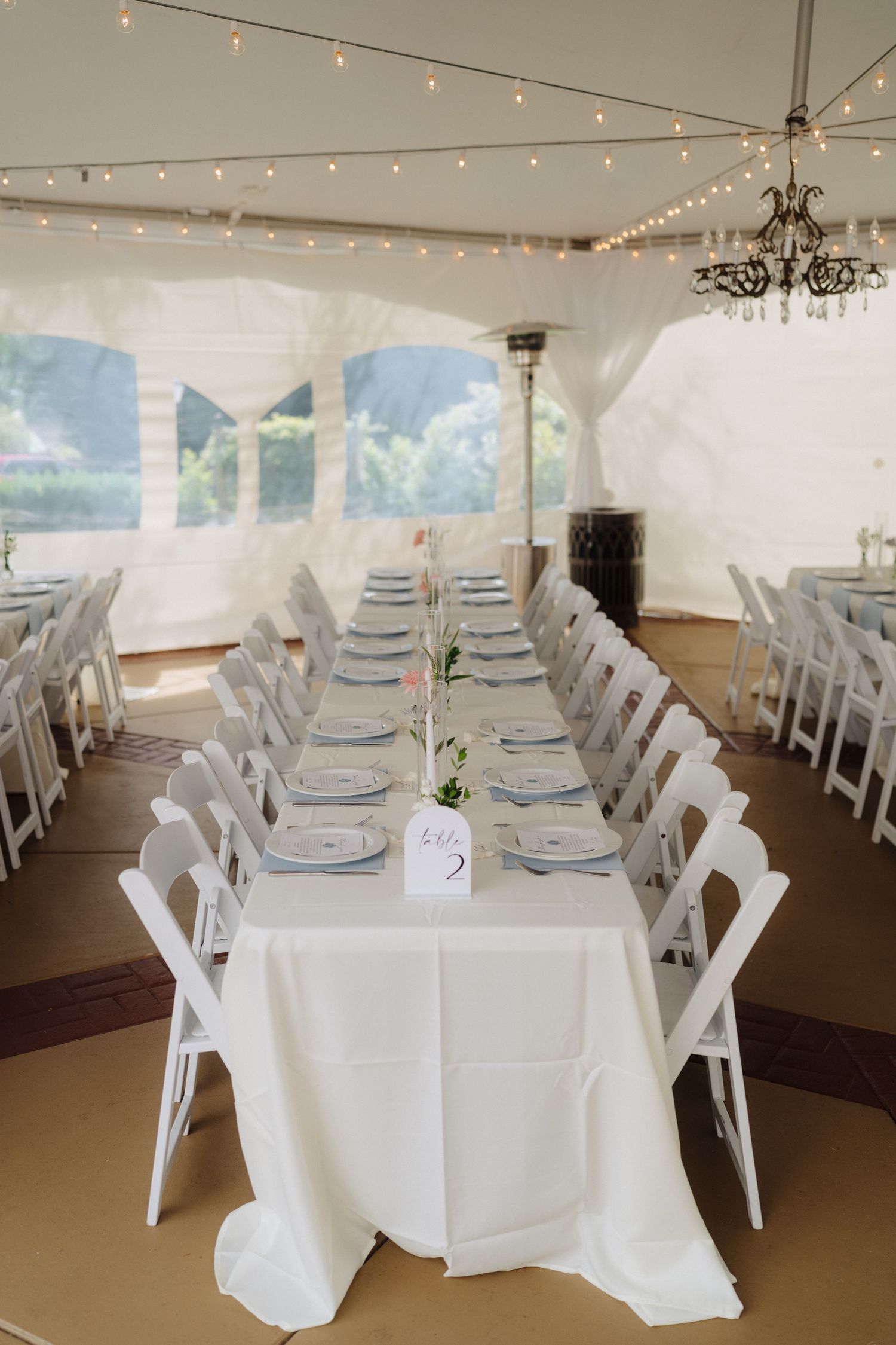 An elegant reception setup with long tables, white chairs, and string lights under a white tent.