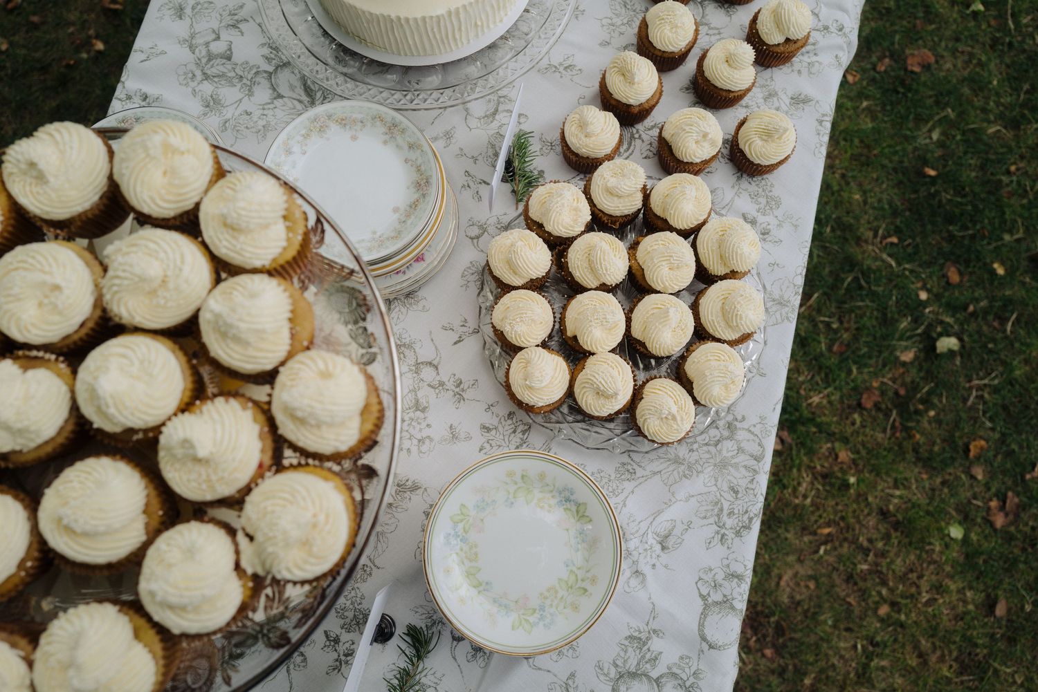 Wedding cupcakes and cake displayed on a white tablecloth outdoors.