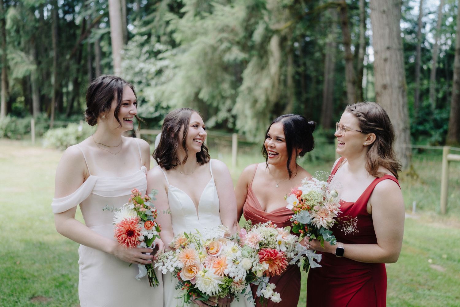 Wedding party members in burgundy and blush dresses share joyful moments in a lush forest setting.