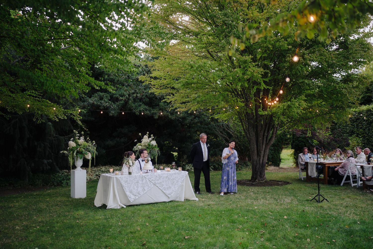 Elegant outdoor wedding reception with string lights, lush trees, and guests dining at tables on a lawn.
