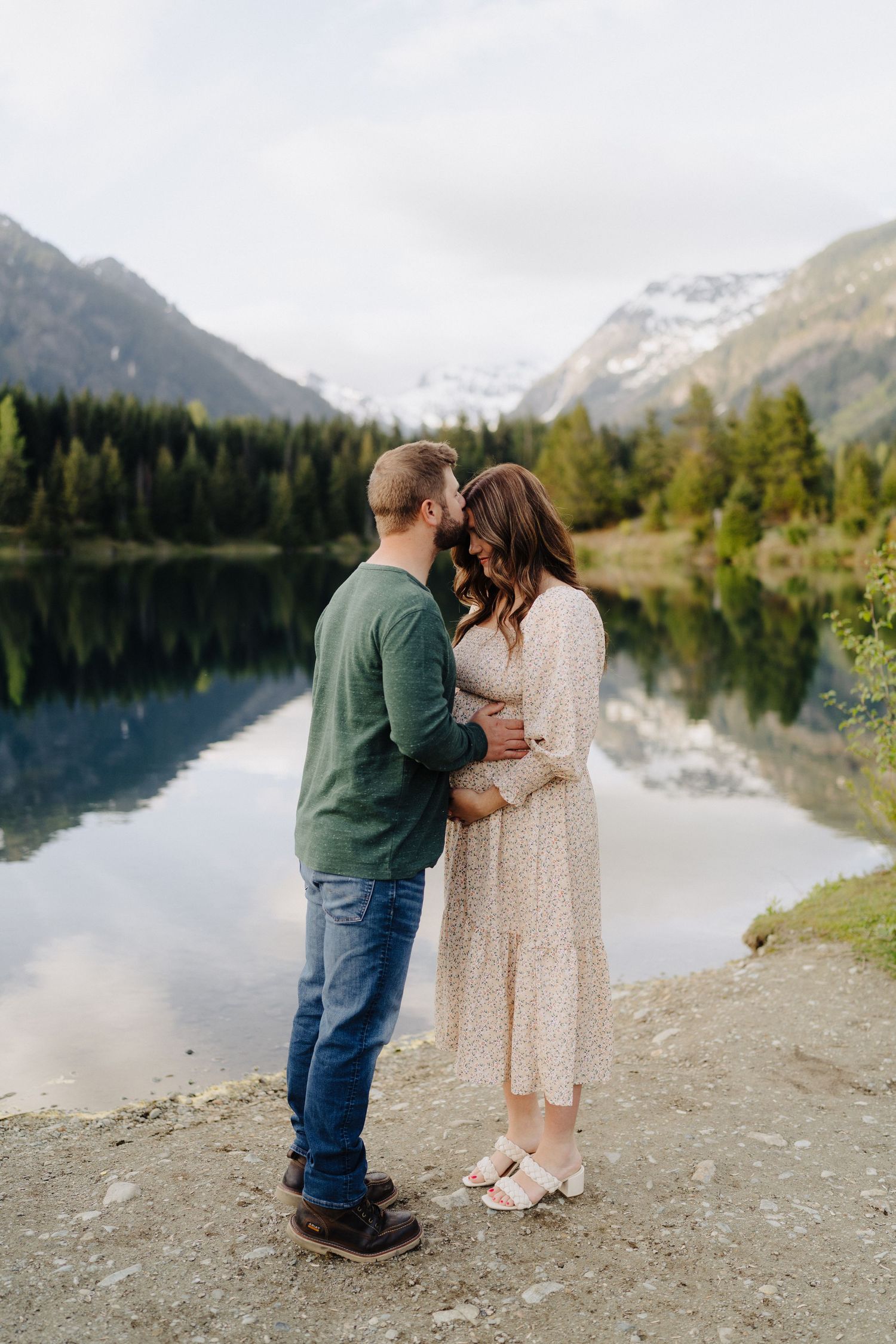 A couple shares an intimate moment by a serene mountain lake surrounded by snow-capped peaks and evergreen forests.