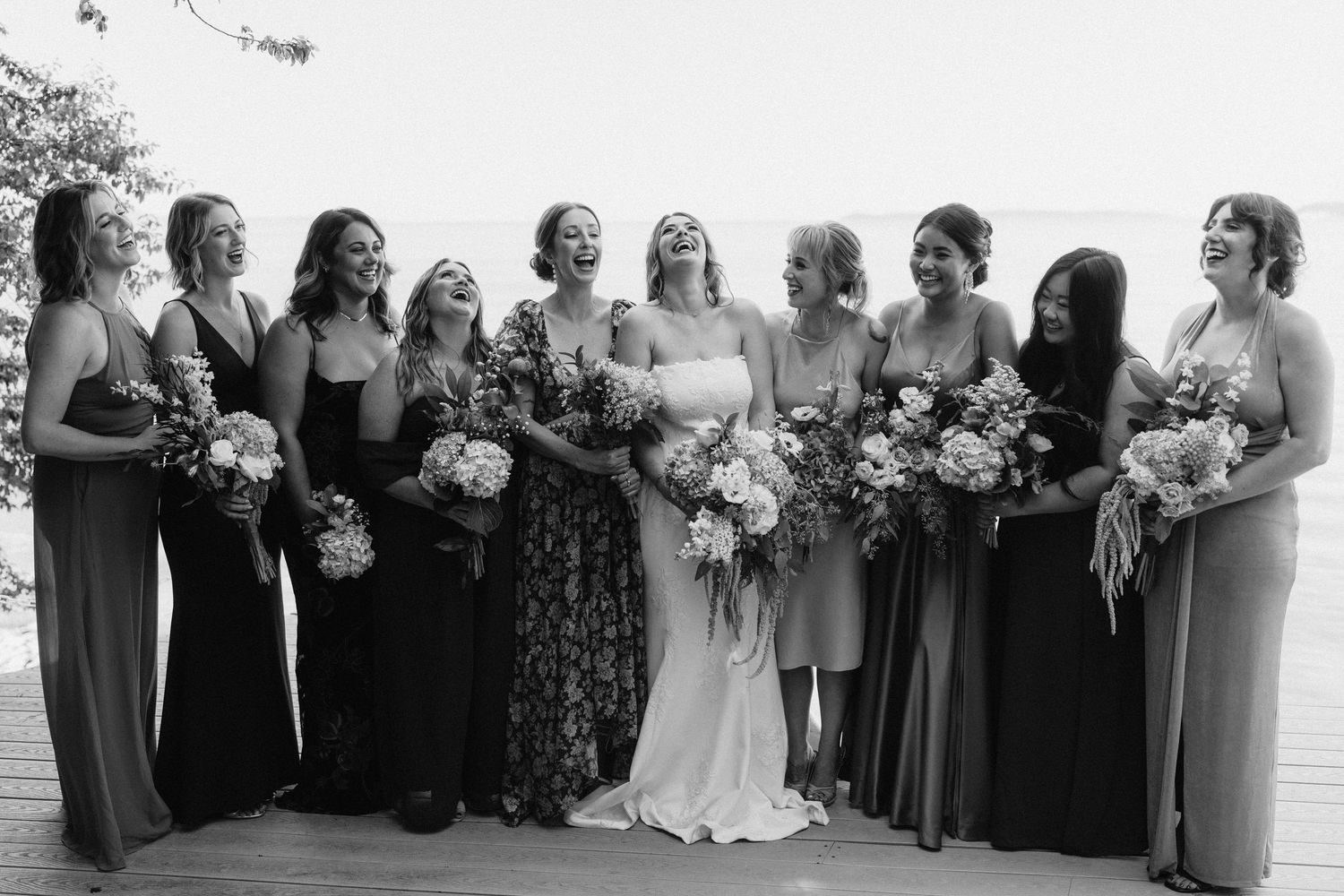 Bridal party in mix-matched formal dresses pose with bouquets on waterfront deck.