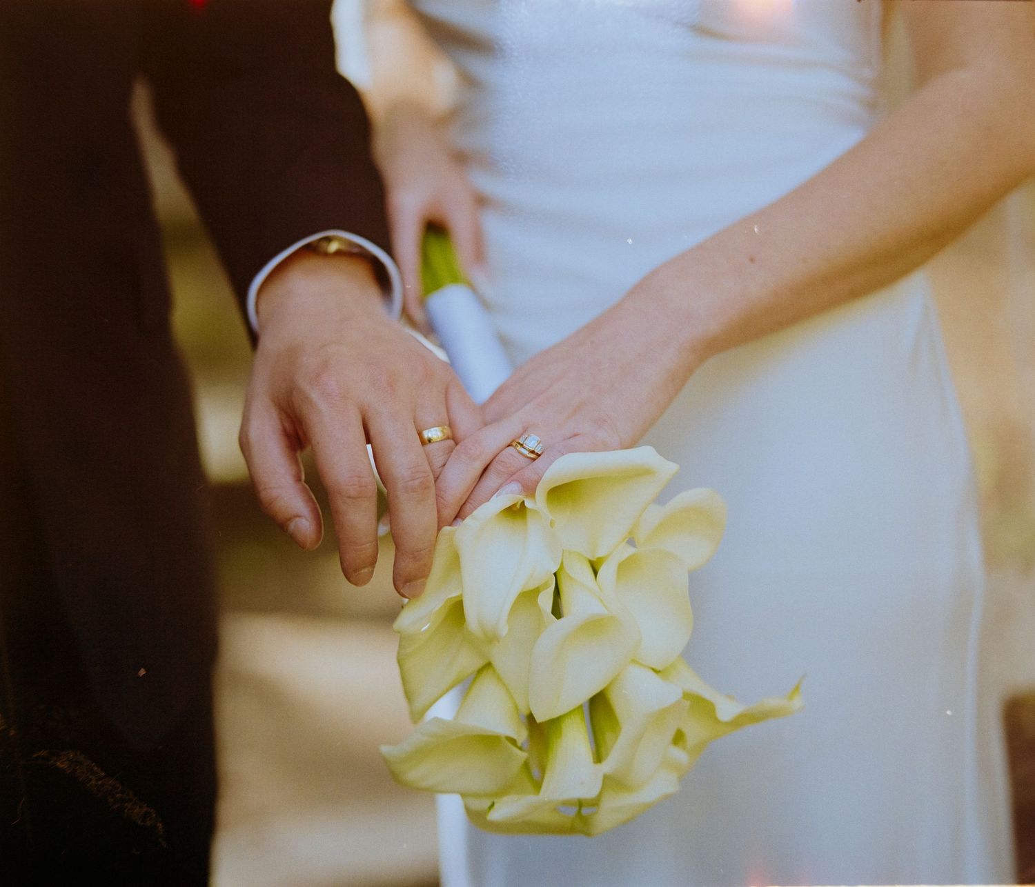Close-up of bride and groom holding white calla lily bouquet, showcasing wedding rings on their intertwined hands.