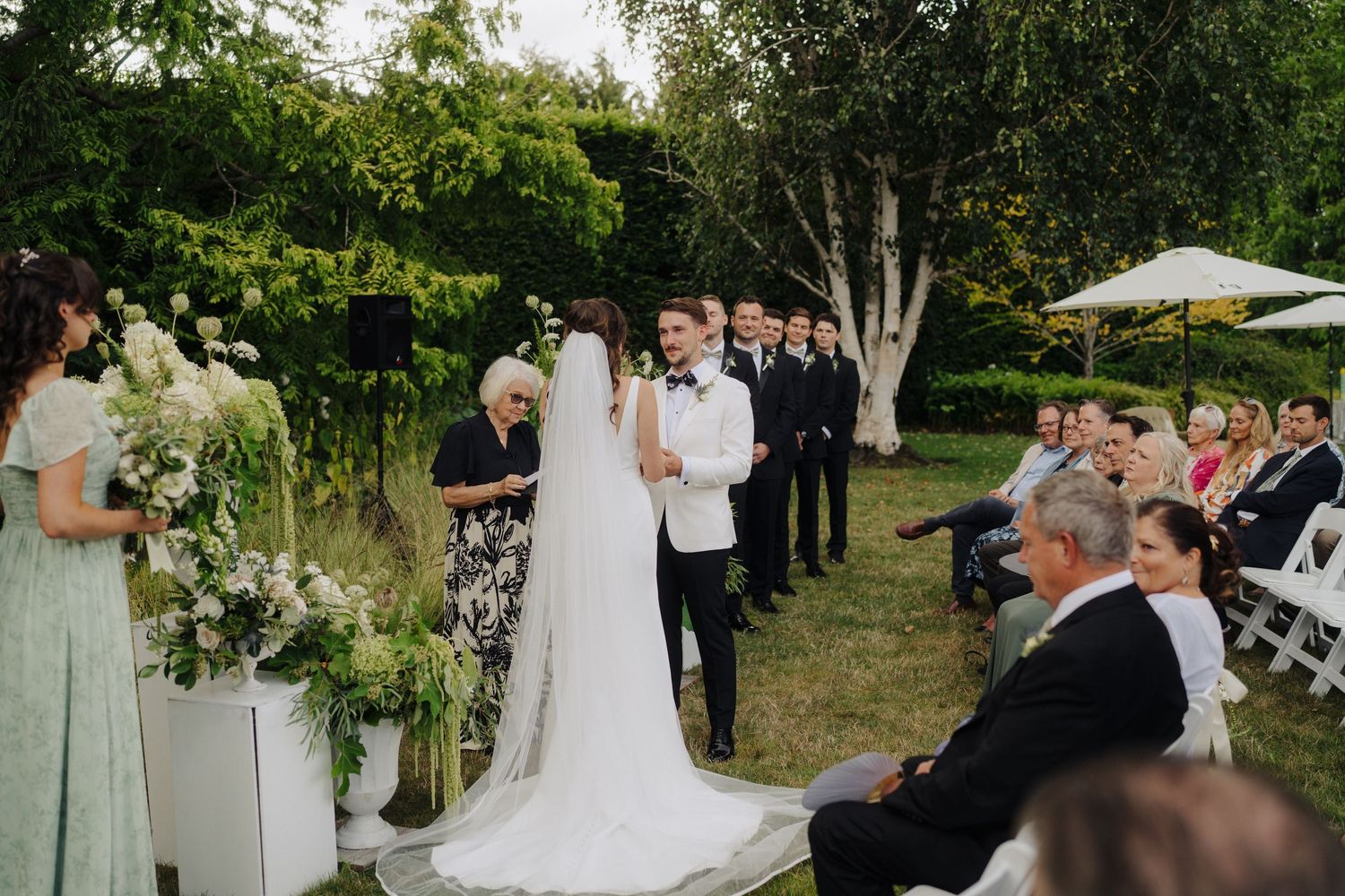 Outdoor garden wedding ceremony with guests seated on white chairs surrounded by greenery.