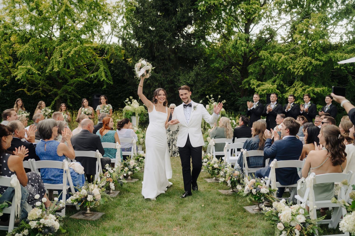 Wedding ceremony setup in a garden with white chairs and guests seated under trees.