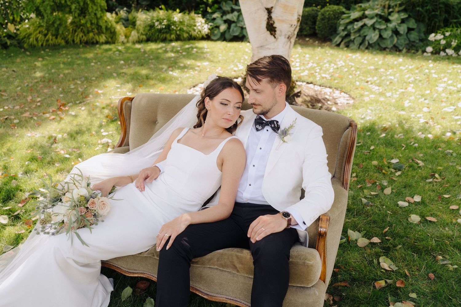 A couple in wedding attire relaxes on a rustic bench in a garden setting.