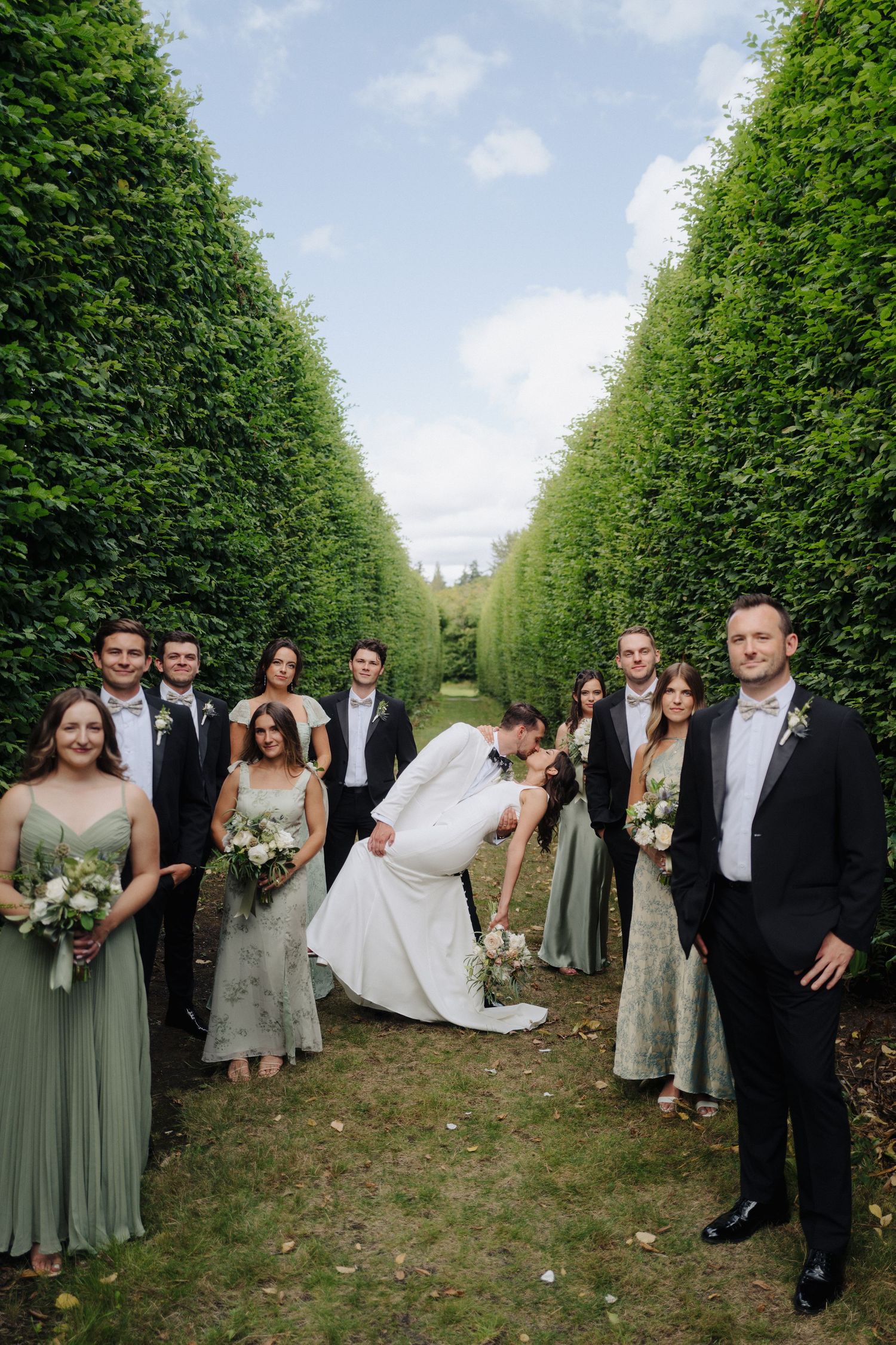Wedding party posing for photos along a manicured hedge garden path.