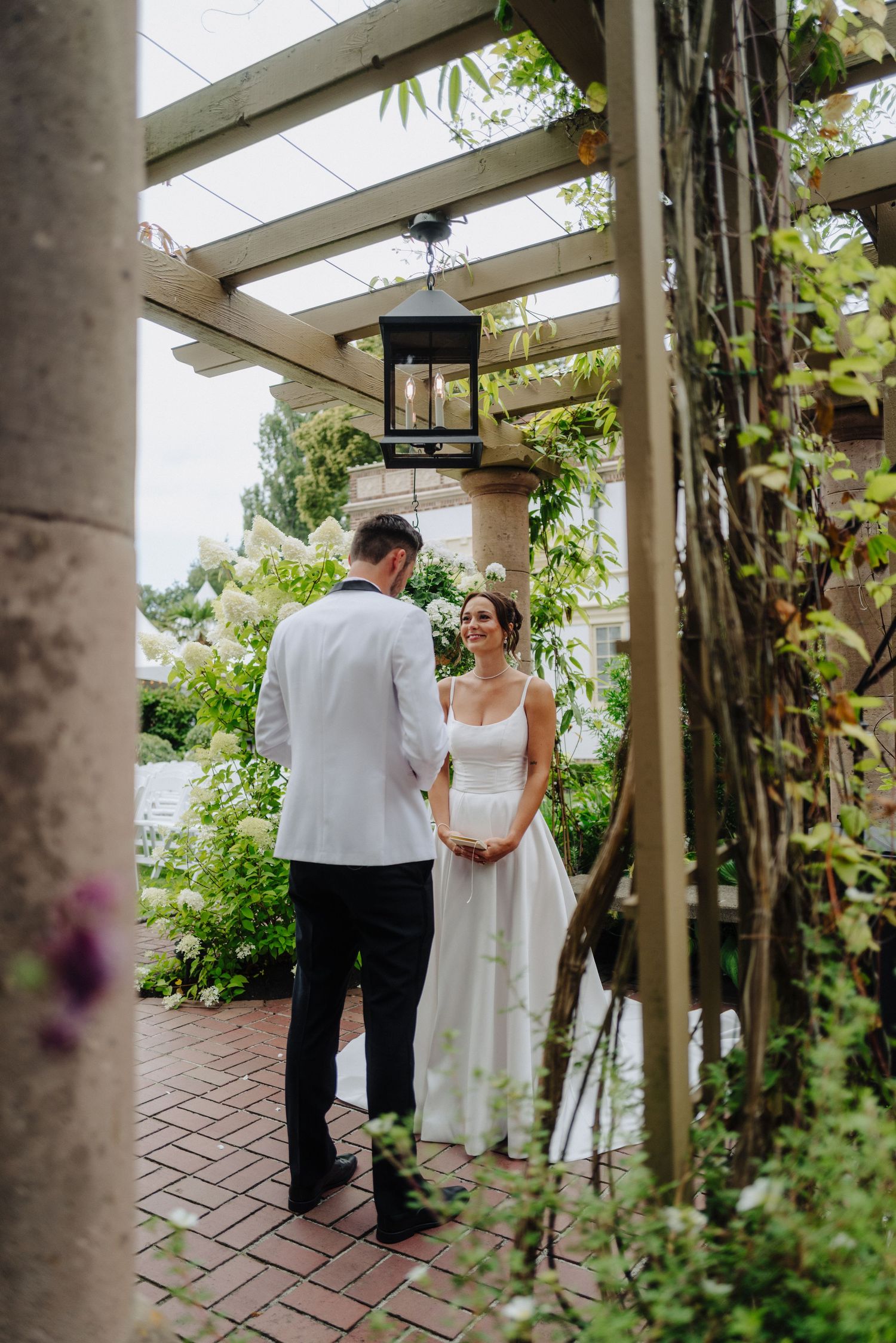 A couple shares an intimate moment in a garden pergola with climbing vines and brick pavement.