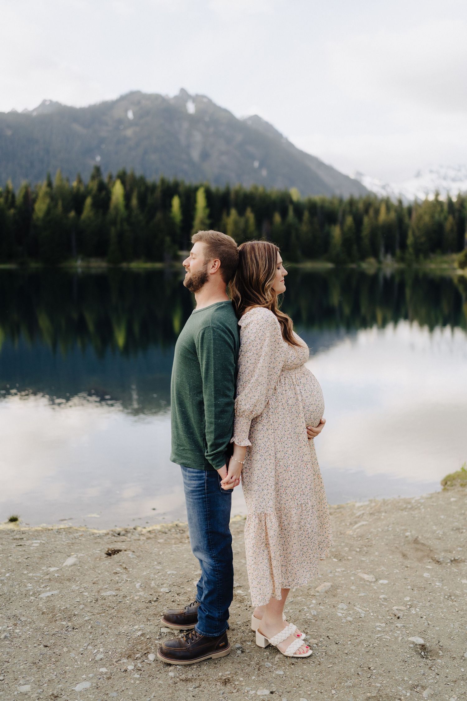 A romantic couple stands back-to-back against a backdrop of mountains and a pristine alpine lake.