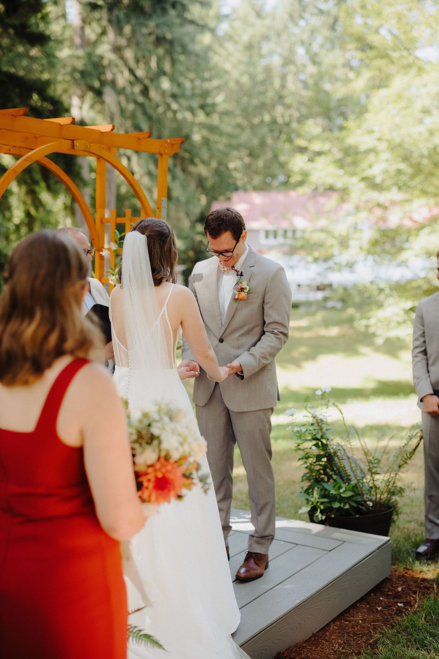 An outdoor wedding ceremony takes place under a wooden arbor with orange and red floral decorations.
