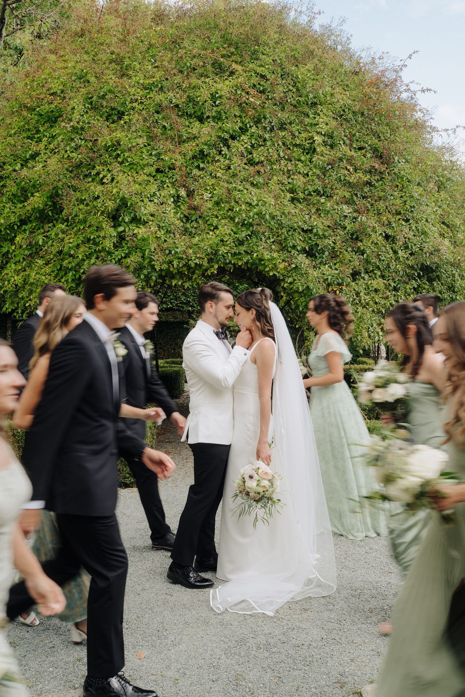 Wedding party members in formal attire moving along a garden path.