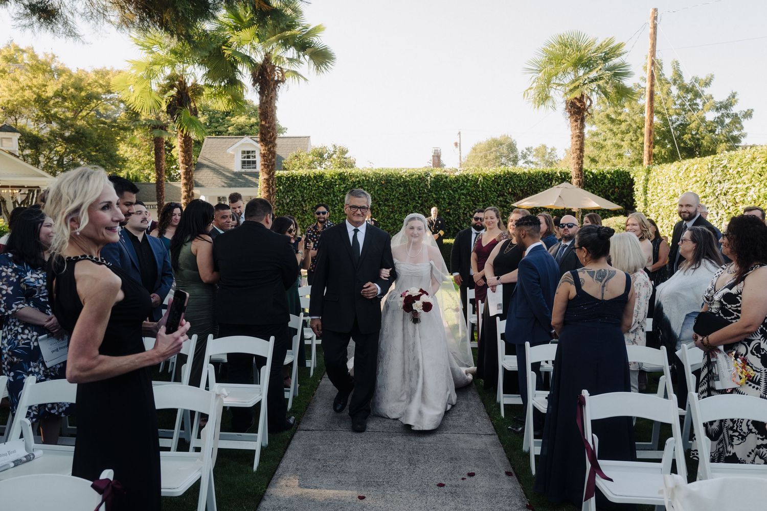 Outdoor wedding ceremony with guests seated in white chairs along a garden path.